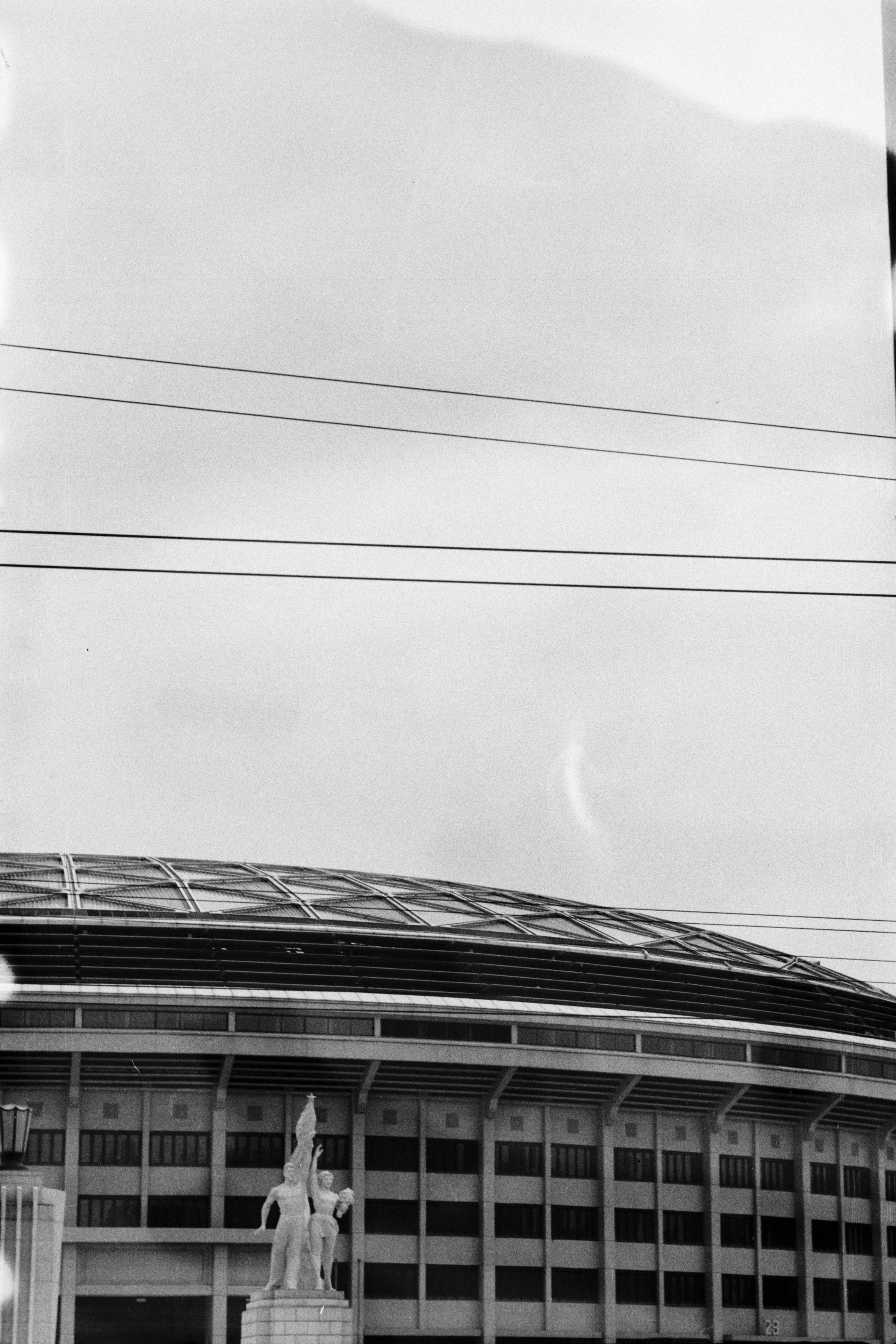 ERA 100 (FF)

English
A black and white photograph of the facade of a large stadium where a communist style statue stands in the front. Power lines can be seen across the sky.

中文
一张黑白照片。照片中一个大型的体育馆外墙。前方有一个共产主义风格的雕塑。天空中有电线穿过。