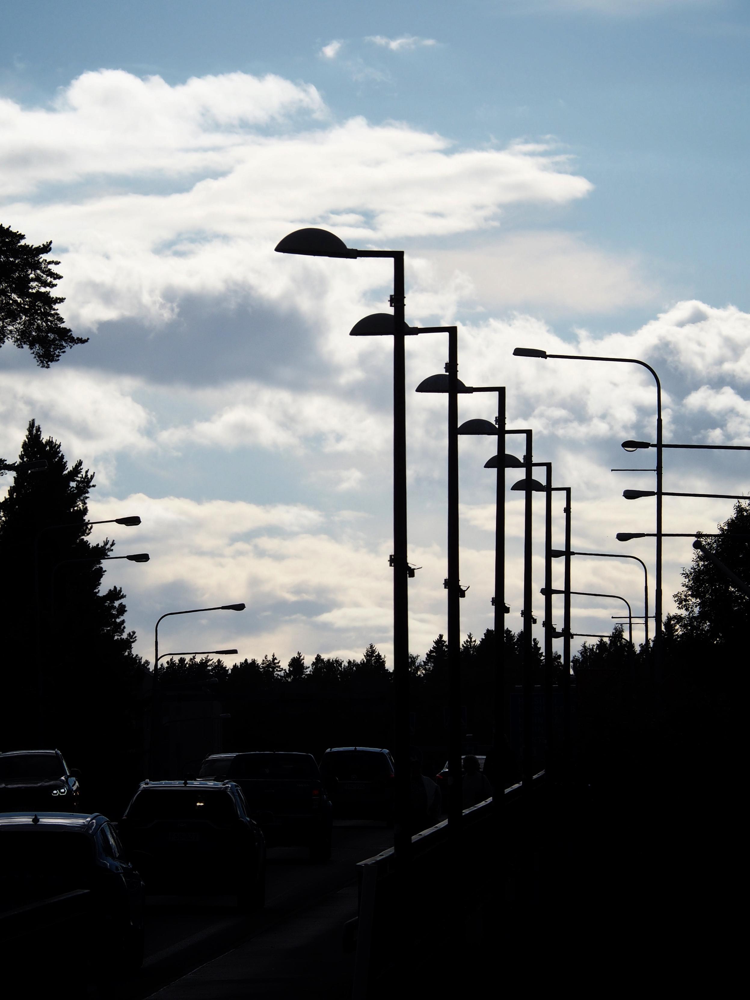 A silhouette of streetlamps along a bridge, with cars passing. The background features a partly cloudy sky and trees.