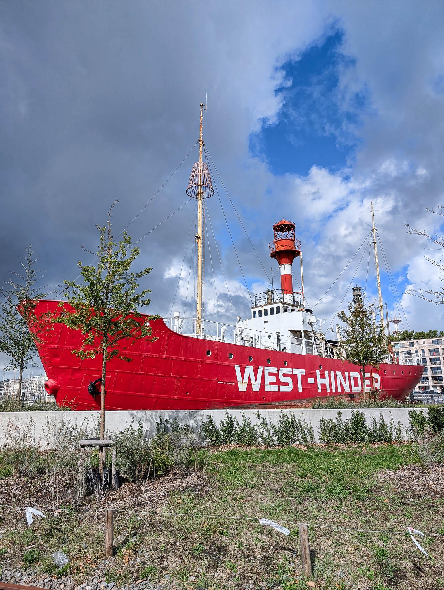 We're in front of a short stretch of dry, coarse land with some patches of dirt and short grass rising gently to a low white wall. Behind the wall there is a ship mounted on display, about forty metres long. The vessel has a red hull and white bridge structure about two thirds back with a red-and-white-hooped, cylindrical, beacon tower emerging from the bridge, topped with a grilled casing in which a a large lightbulb sits. The words "West-Hinder" are in bold white on the ship's hull. A thin, pale-coloured mast also sits forward of the bridge with a flared, basket-like cage near its top. There's a sky of cloud threatening rain with large patches of blue overhead and a few short, thin trees with light foliage are dotted around this rather industrial location.