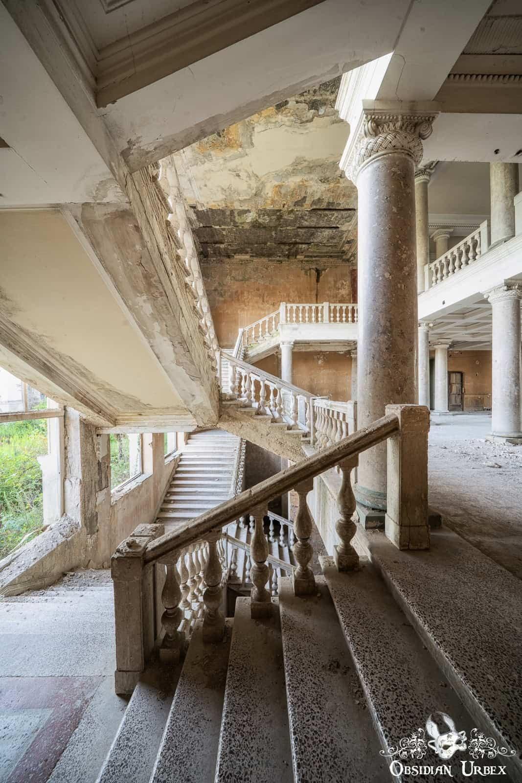 A vast, decaying interior showcases a grand multi-level staircase with ornate balustrades and large columns, reflecting the building's abandoned state.