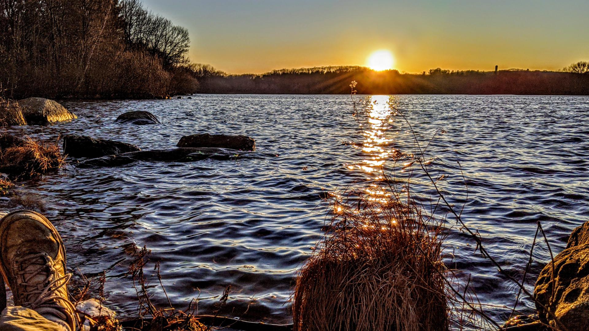 A deep blue sky with puffy white clouds partially silhouetted by the sun, which is setting behind a low wooded ridge on the far shore of a lake. A soft breeze is gently rippling the lake, creating a long wavy streak of golden orange reflecting the sun. That streak ends in a small patch of water grasses a few feet offshore, but the nearest ripples catch shifting blobs of that golden light. We are sitting on a flattish gray rock slightly curved in front at the near edge of the lake.