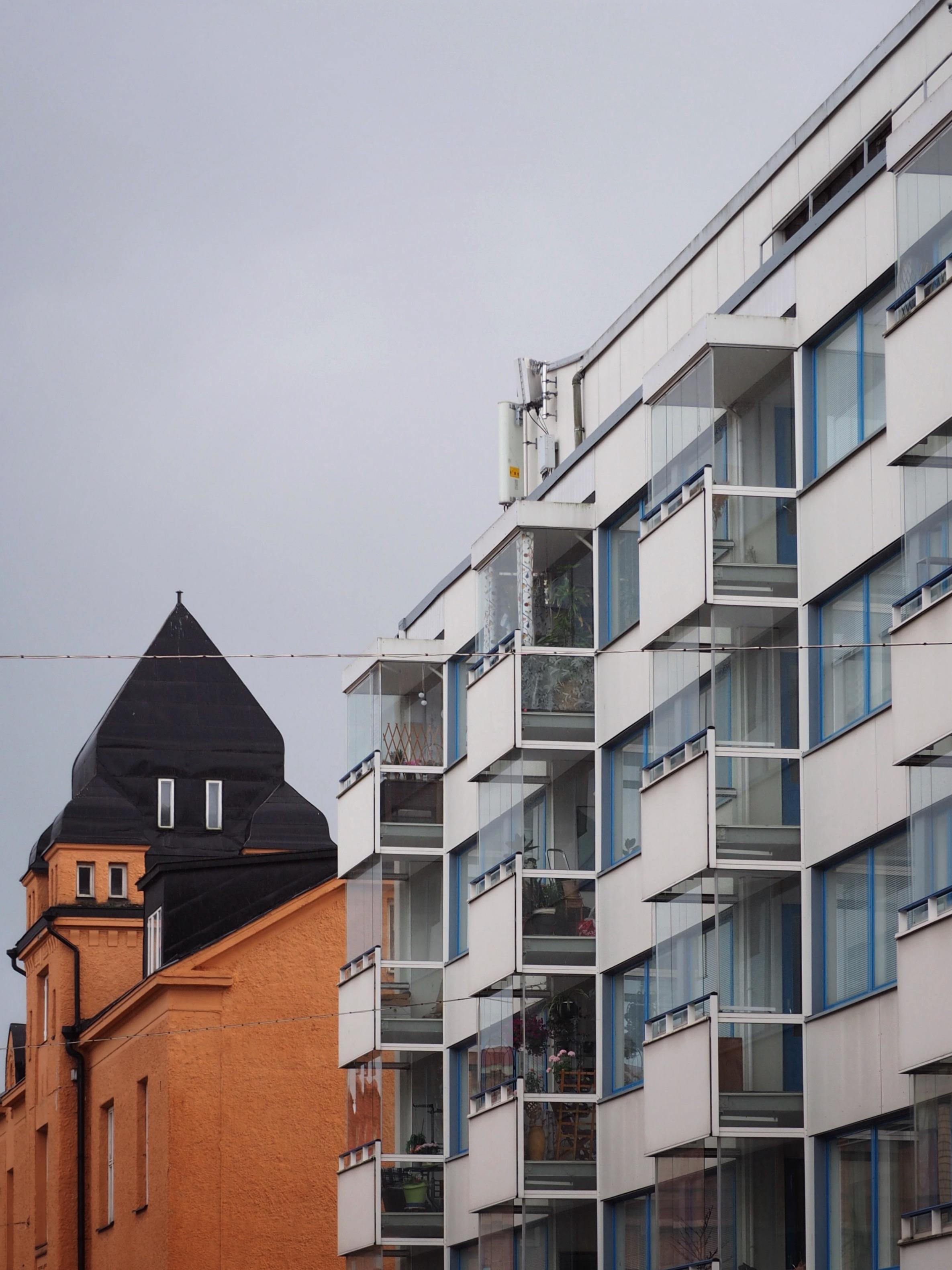 The image shows a contrast between two buildings: a modern white apartment building with balconies on the right and a traditional orange building with a black peaked roof on the left, under a gray sky.