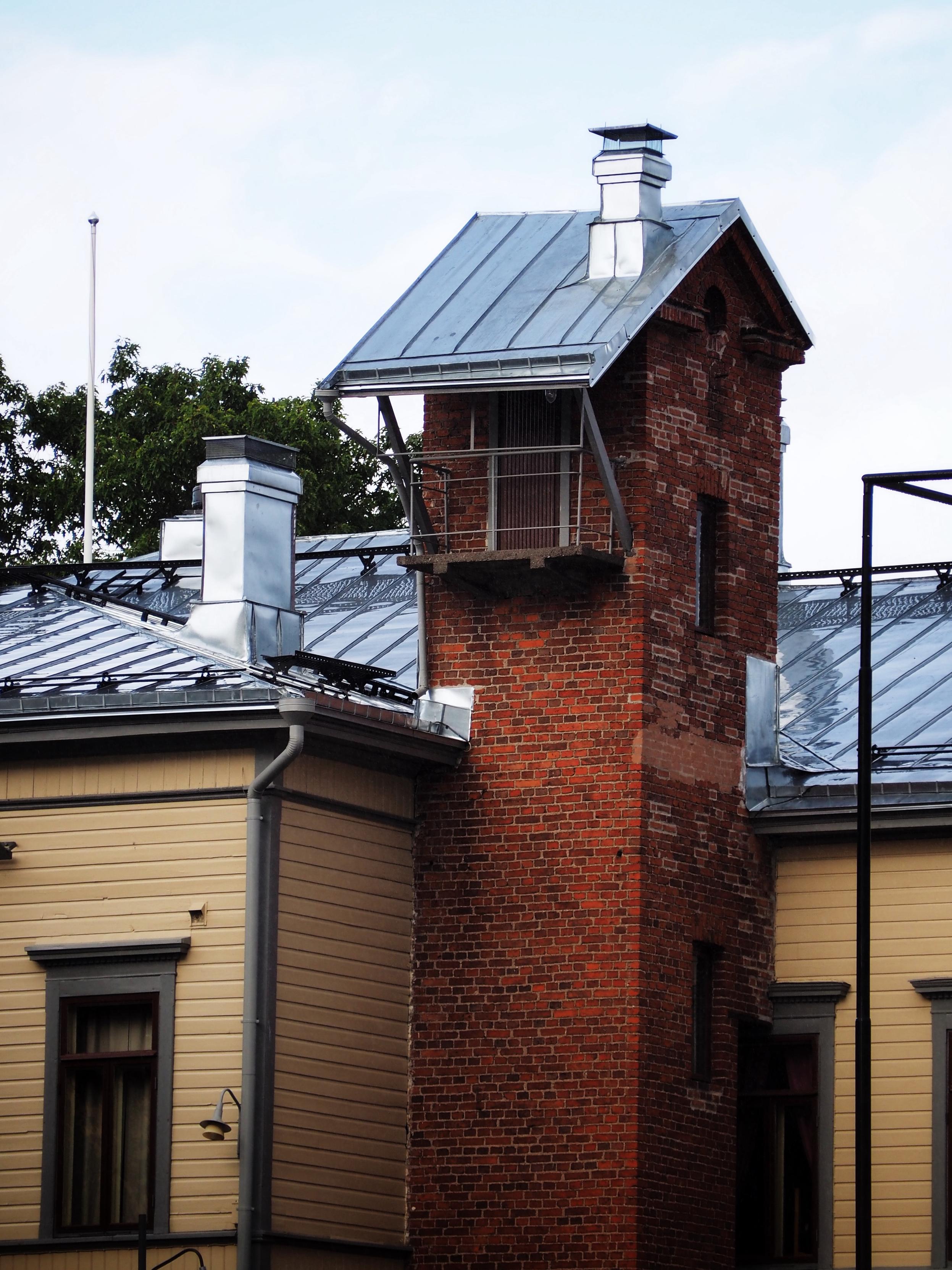 A close-up of a building featuring a brick tower with a sloped metal roof and a small balcony. The structure has a combination of red brick and wooden elements, with visible windows and a clear sky in the background.