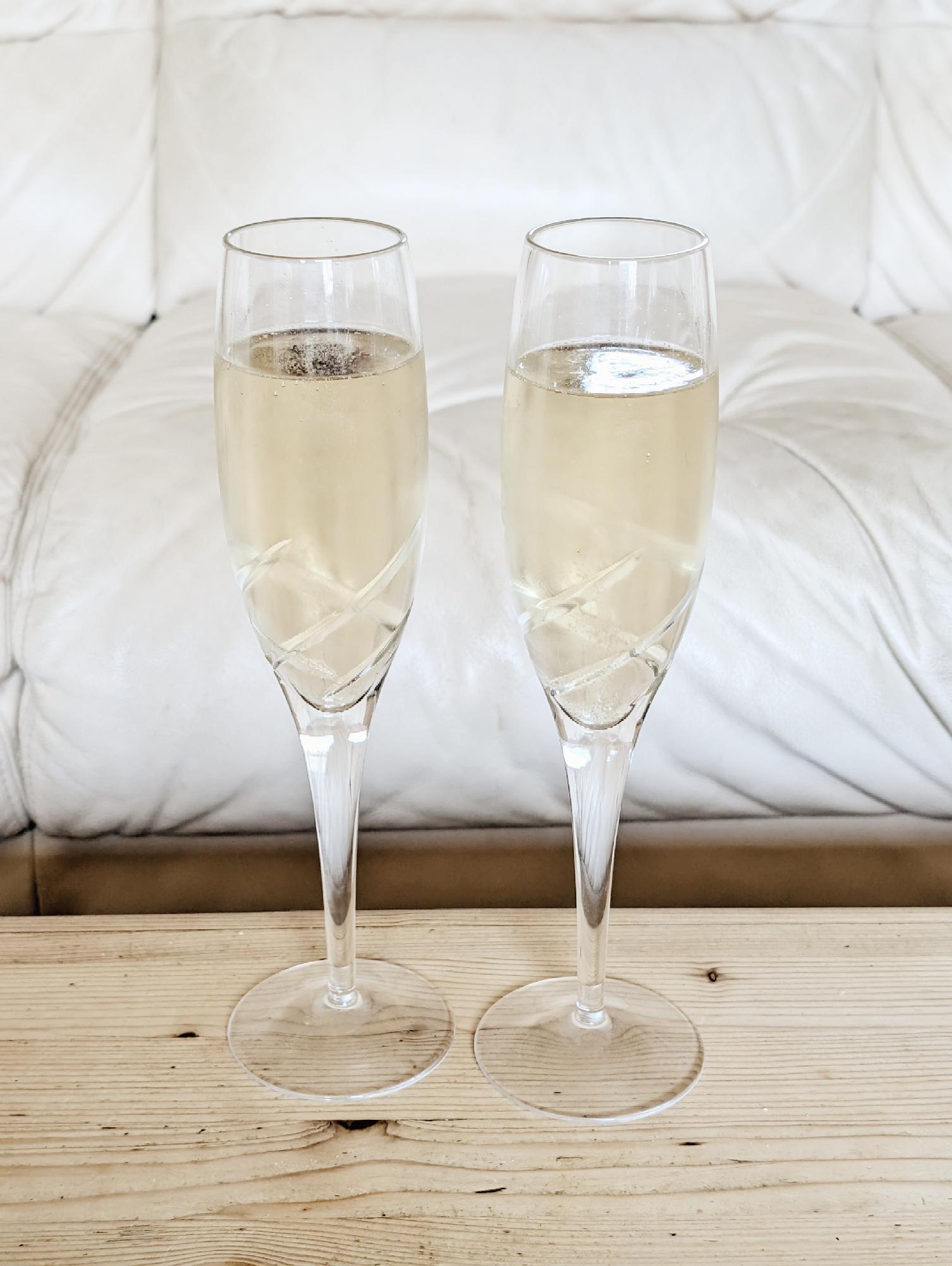 A close view of two champagne flutes with champagne on a pine wooden chest surface in front of a cream leather sofa. The scene is illuminated by natural daylight and there's the reflection from a window in the surface of the champagne flute on the right.