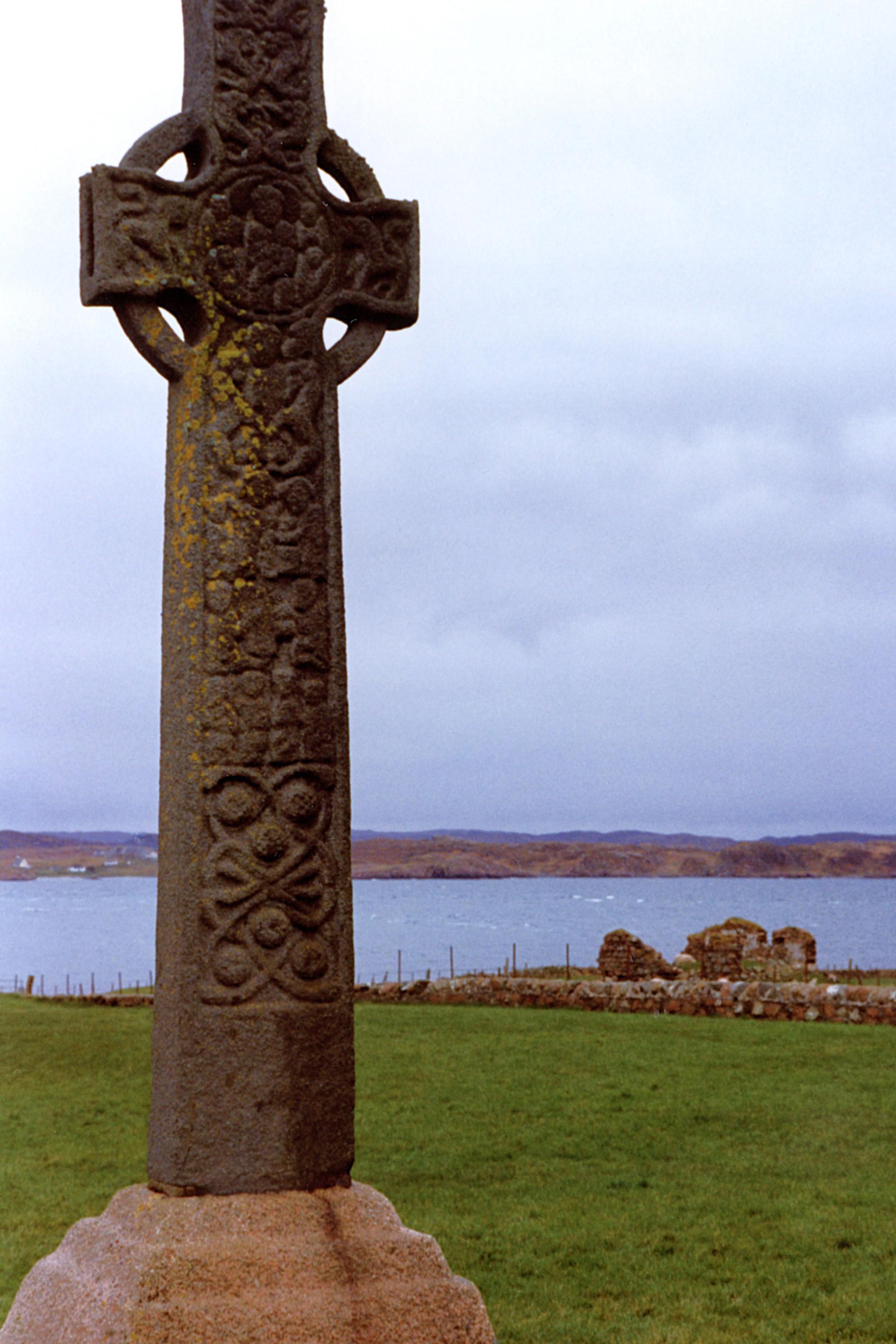 A large, stone, Celtic cross stands to the left of the frame, with grass and water behind.