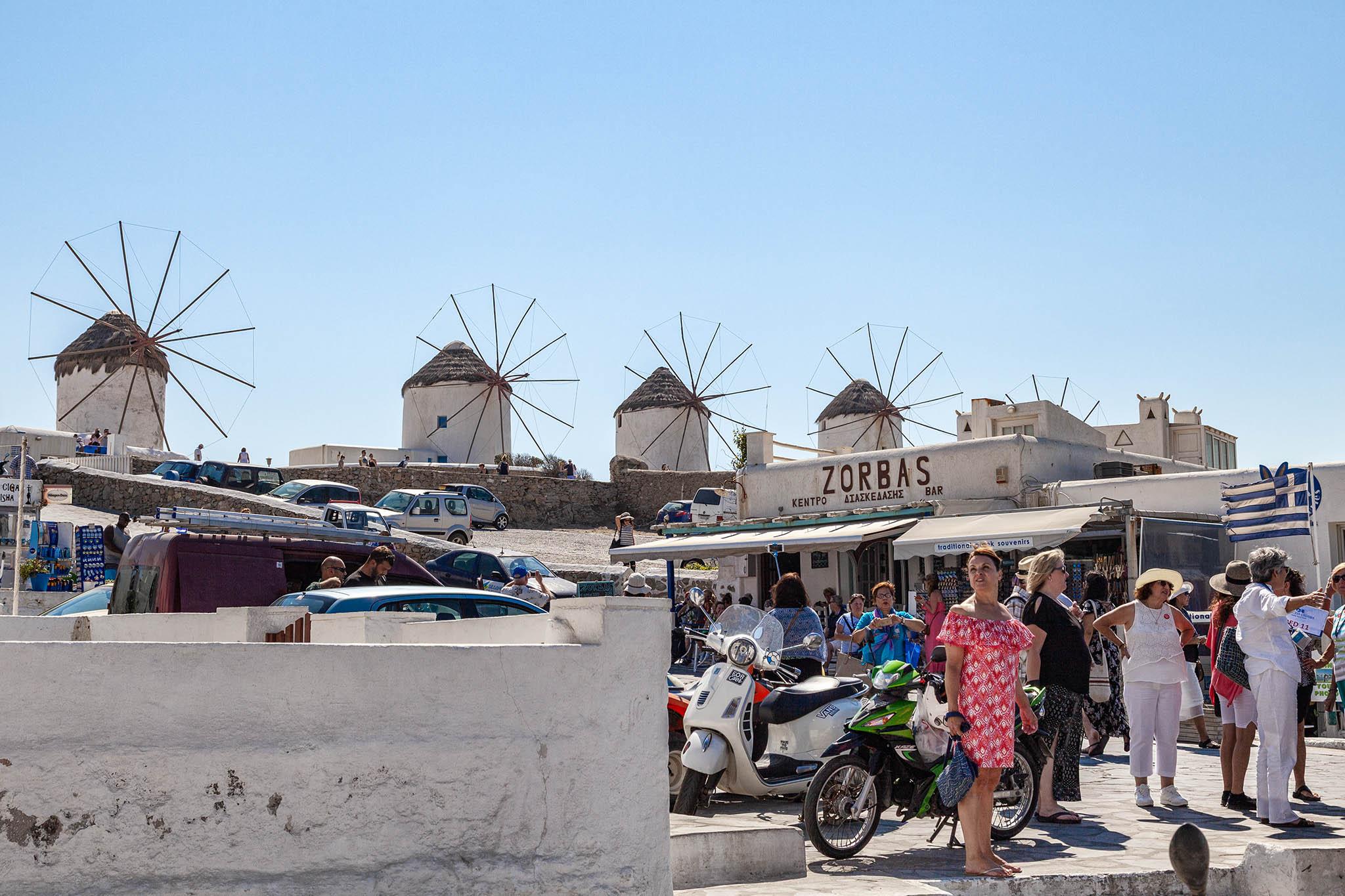 There's not a cloud in the piercingly blue sky on this hot day. There are low buildings and walls around a sloping bit of stone-paved land, all of them painted and plastered in white, typical of Greek structures in the Mediterranean. Tourists gather near a wide opening in the nearest wall on the right and their guide holds a Greek flag on a pole to help them keep track of his position. Motorbikes and cars are parked on the sloping area behind the wall and there's a bar with some awnings stretched out in front just behind the tourists. The name "Zorba's" is written in large black lettering on the upper part of the low building. Just beyond another low wall that runs around the top of the sloped parking area we can see five small windmills, one of them largely hidden by some other buildings. Each windmill is cylindrical, with thick white walls and a thatched, conical roof. Twelve, long, thin poles form the shape of the windmill blades but there are no sails mounted between them, leaving 