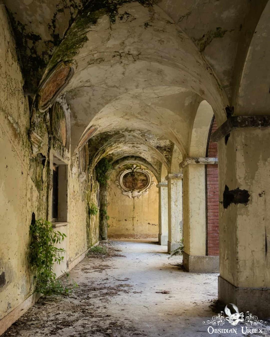 The decaying cloister of an abandoned 17th-century Italian convent, featuring arched ceilings, overgrown moss, and faded wall art.