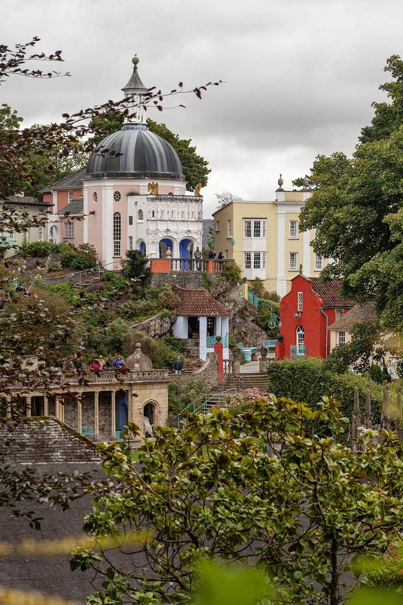 A portrait orientation shot taken on a dry but overcast day. We're looking at buildings in an Italian riviera style tucked into a woodland setting, framed by trees either side and in front of us. Some buildings are colourful and one large structure is topped with a dome and small clock tower on its apex.