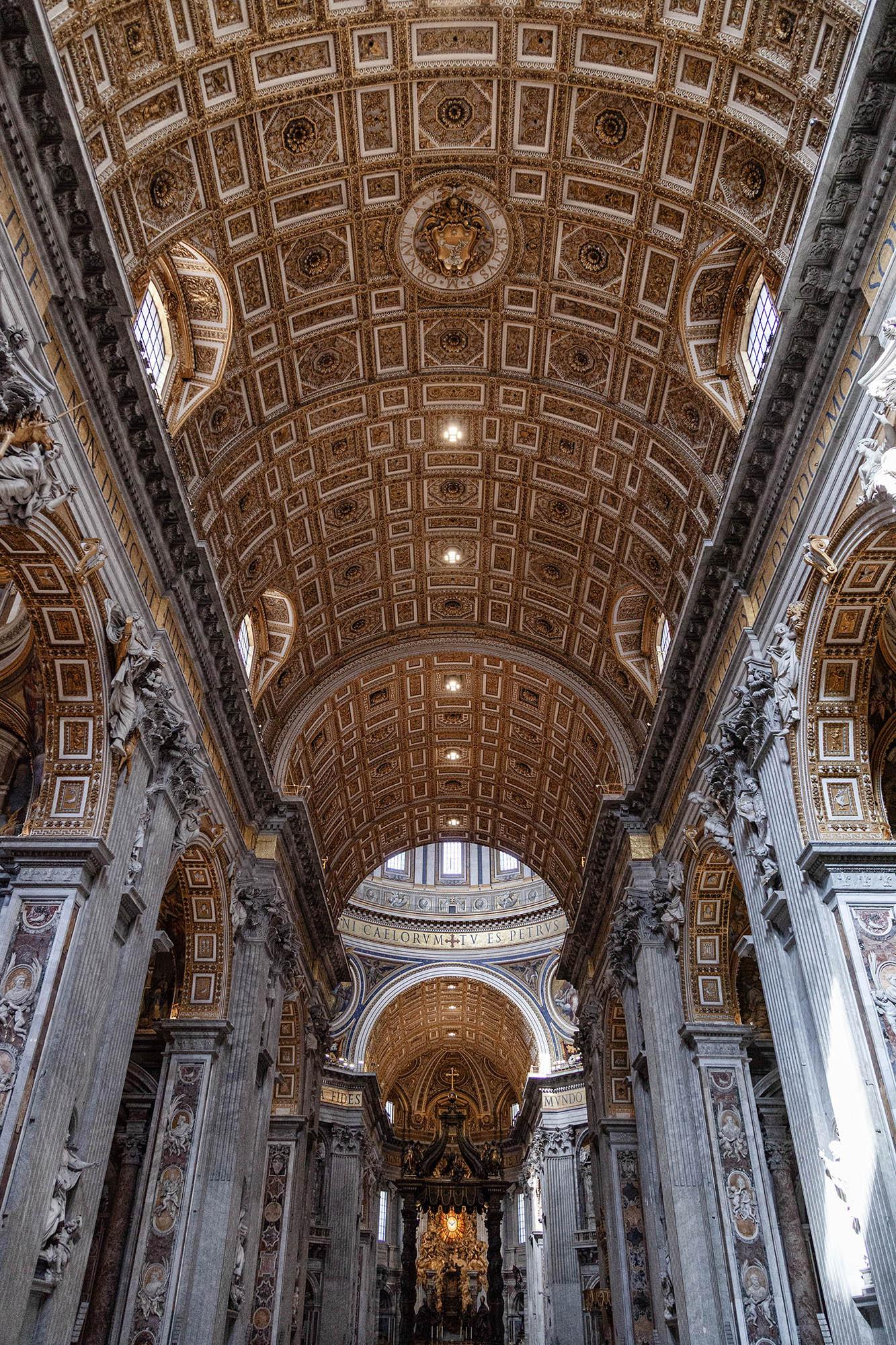 A portrait orientation view shows the inside of St Peter's Basilica in the Vatican City. Huge, square pillars are topped by arches in two rows stretching away from us. Between them the ceiling forms a curve until it reaches the far end where a domed opening can just be seen and daylight comes in from above. The ceiling is highly decorated in panels but the height of the church makes it difficult to make out any details.