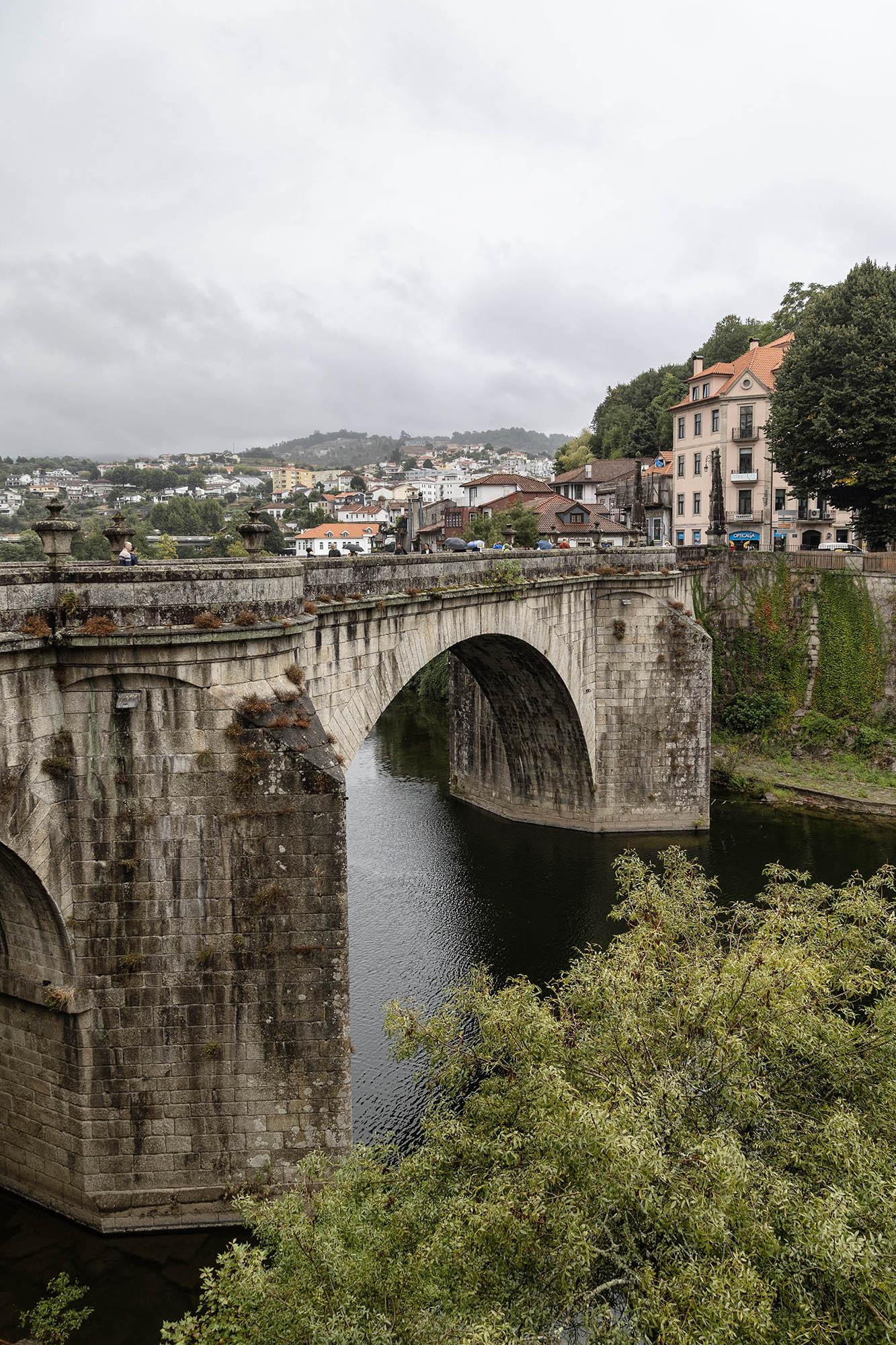 We're on a high bank on one side of a river on an overcast day. Both sides of the banks are steep, almost sheer, and covered with greenery on their sides and topped with bushes and trees. A bridge spans the river. It is wide and made from pale stone, dirty with exposure to the elements, with two thick supports connected by a long, shallow arch. On the far bank, back from the bridge an old, multi-storey building rises and the roofs of a village slope down in the distance beyond.