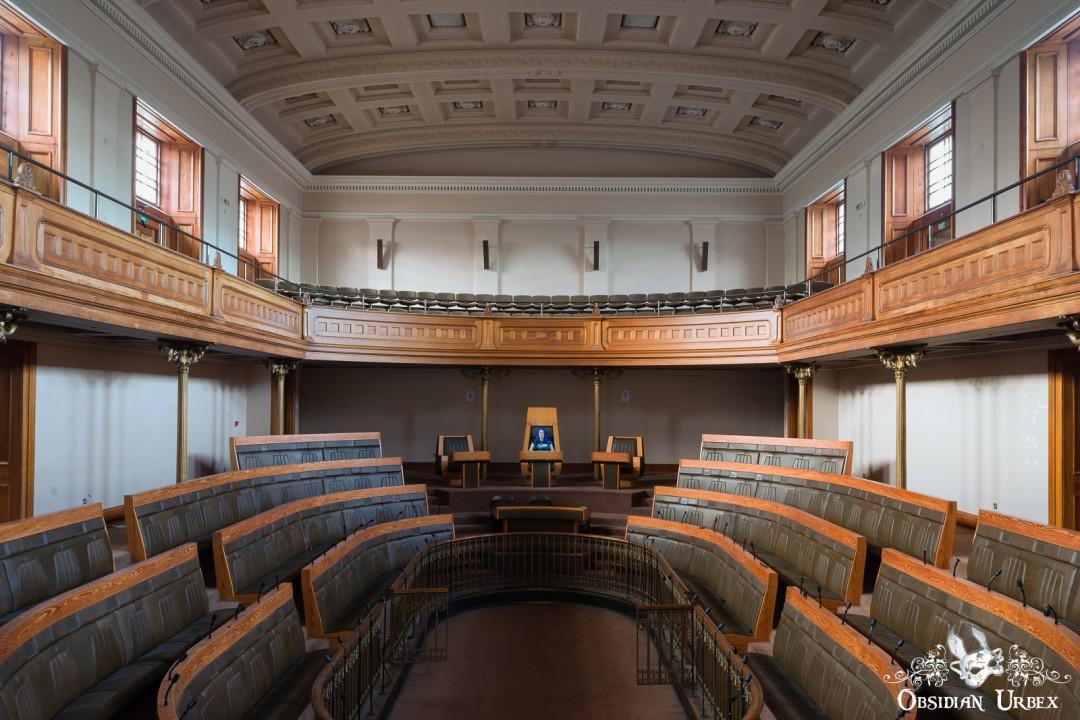 An empty, ornate assembly hall features tiered wooden seating and a raised platform with a portrait. A balcony lines the upper walls and a detailed ceiling overhead. A girl with blue hair sits in a chair at the far side of the room