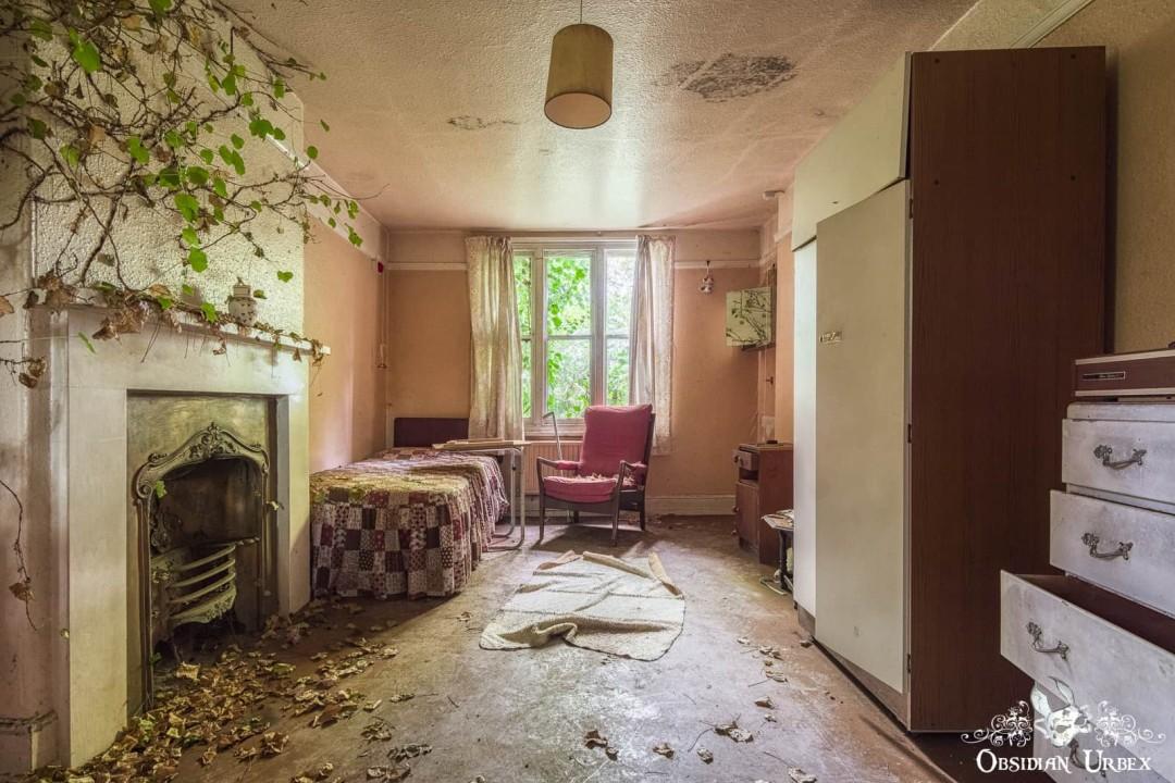 An abandoned small room in an English care home, with vines over the fireplace and leaves on the floor. Beds, wardrobes, and a red armchair furnish the space.