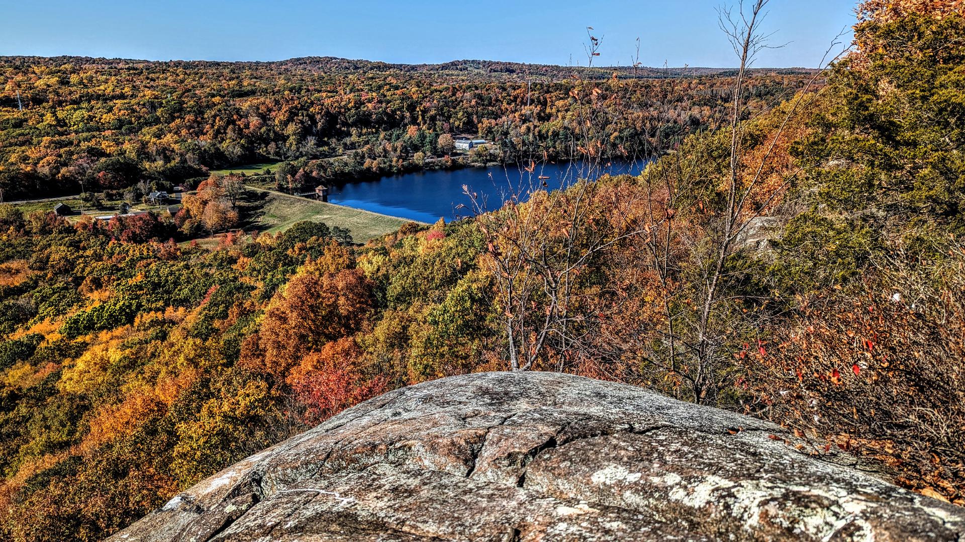 A cloudless sky of robin's egg blue. Sitting on an outcrop of light reddish-brown rock liberally covered with light gray lichen. The outcrop looks out over a portion of a long, narrow valley a few hundred feet below and a long, flattish ridge across the valley. A rural two-lane road through the valley. The grass-covered earthen dam and lower end of a reservoir is to the right of our pic. The water is deep blue on this perfect autumn day. There are wide swaths of mowed grass and pastures near the dam and a few farm buildings and houses visible, but the vast majority of the valley and the ridges on either side is covered with a forest sporting slightly past peak but still vibrant foliage of golds, yellows, oranges and brick reds.