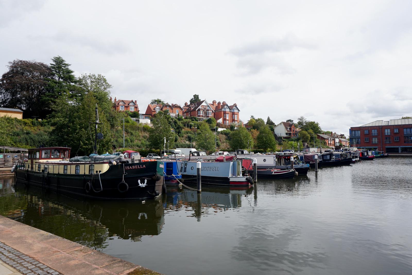 several narrowboats moored in the basin, all in a line. What appears to be an office building at the far end.
Behind where they are moored there is a hedge running into the distance and several large houses can be seen on top of a hill in the distance