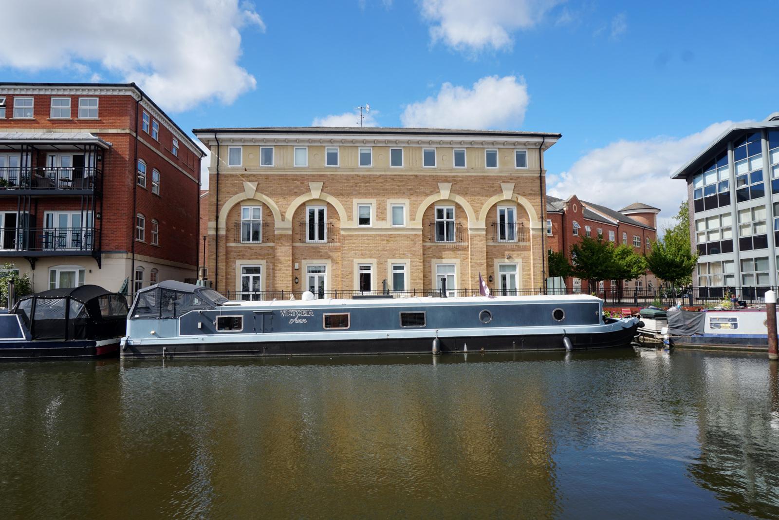 A rather long, quite modern looking grey canal boat moored at the far side of the basin in front of a building, possibly apartments built with light brown bricks  with cream surrounds to the windows