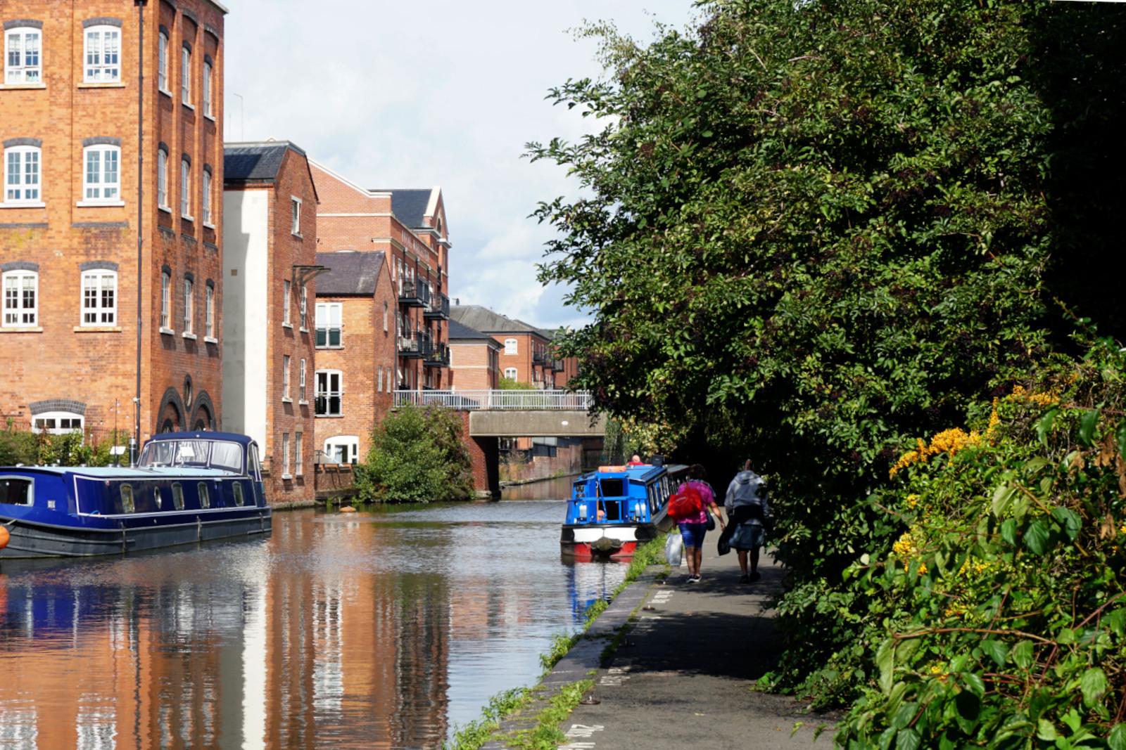 A canal , the towpath on the right has 2 people walking along it, overhanging trees and bushes and a canal boat moored just before a road bridge crosses it.
The opposite bank has modern 3-4 storey buildings and a larger canal boat moored.