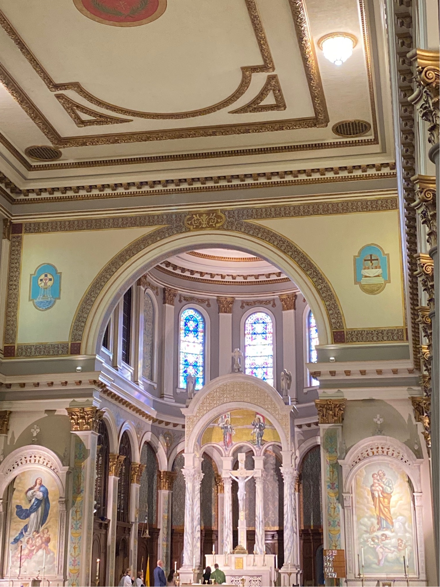 Color image, interior, Joan of Arc Roman Catholic Church, Indianapolis, Indiana, USA, circa 1920. Image shows the area beyond the altar, the structures of the altar, and the stained glass windows above the altar, all of which are arc forms, with the church walls adorned with mosaic images, sun streaming through the upper level stained glass windows.