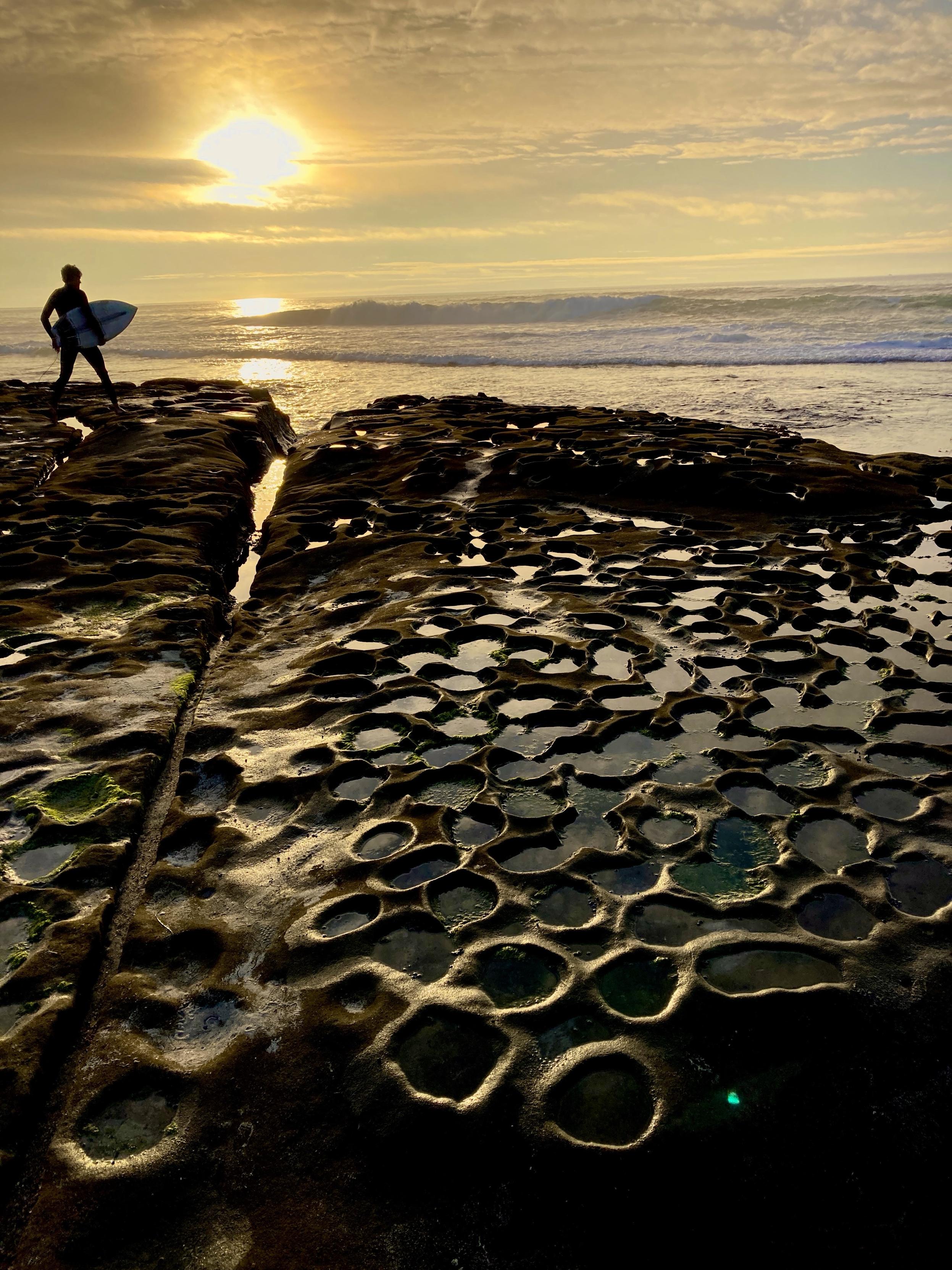 A silhouette of a surfer standing on rocky terrain during sunset, with tide pools reflecting the golden light. The ocean waves are visible in the background.