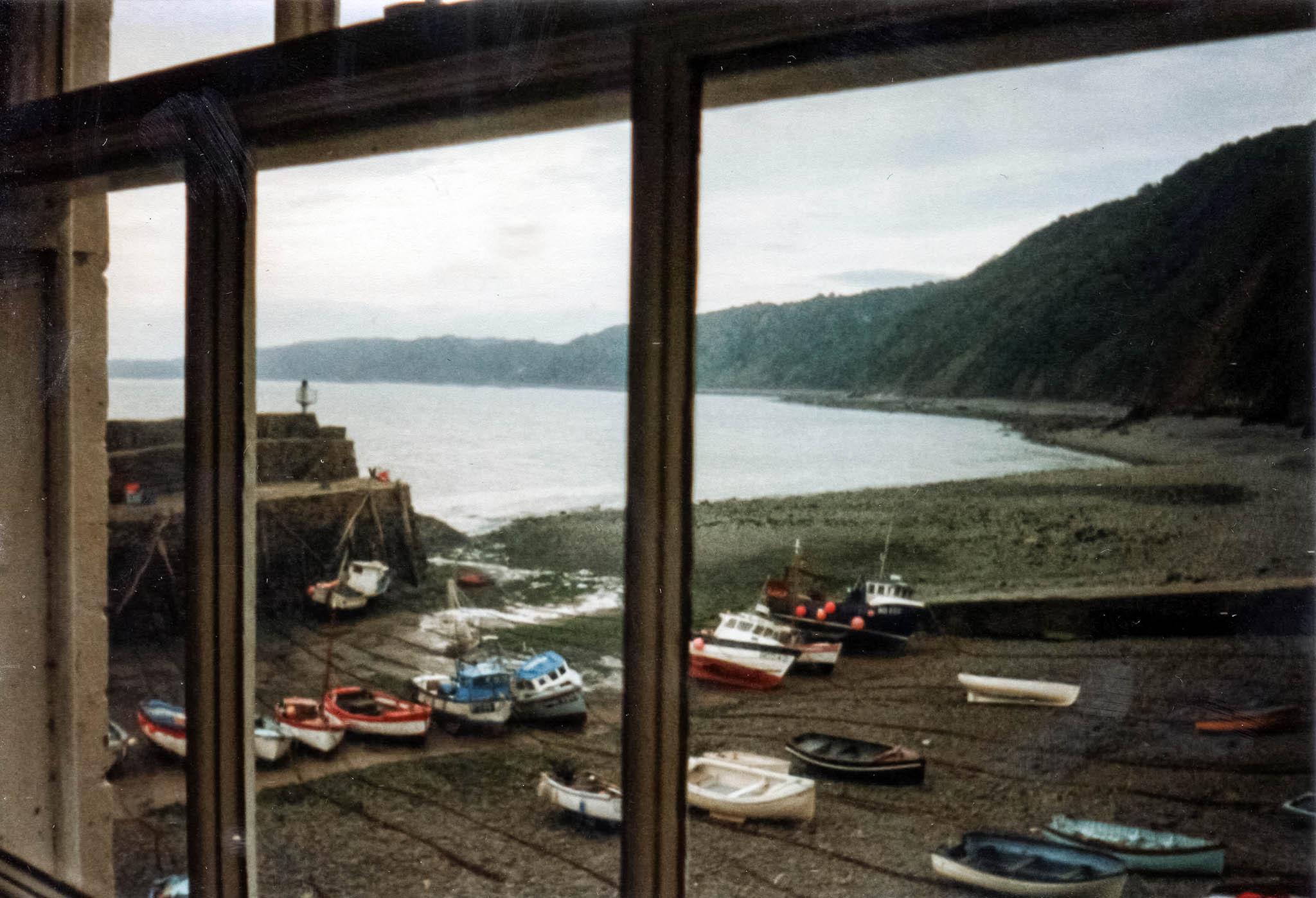 Scan of a colour photograph. We're looking through a window out over a bay. The window has no net curtains up and is a single-glazed, sash window type with each half of the sliding panels split into three panes, the woodwork painted cream but flaking and dirty and showing age. Outside it's an overcast day with a sense of dampness in the air that's not quite threatening rain. The bay is shallow and the tide is out so there are over a dozen small boats on the damp shingle, leaning and waiting for water to come in and float them. A high sea wall is on the left but the bay is mostly exposed directly ahead and to the right where a hilly headland wraps around the bay under the grey sky.
