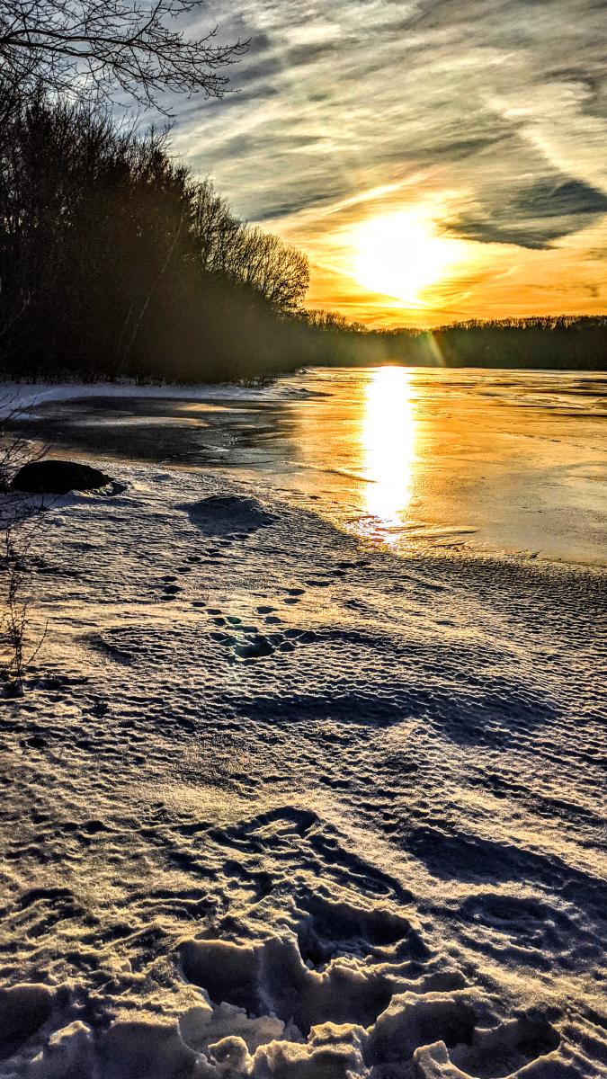 Standing near the shore of a frozen lake. The ground around us is coated with an inch or two of snow that has melted and refroze, capturing small animal tracks and other details in the ice. The sun is setting over the wooded far shoreline, casting deep shadows across our miniature tundra. Leafless trees in the forest surrounding the lake are silhouetted in the light. The sky is filled with streaks of hazy thin clouds, tinted gold and orange along the horizon. The sun is an enlarged blob of bright yellow behind the veil of haze, creating an elongated bright reflection on the ice.