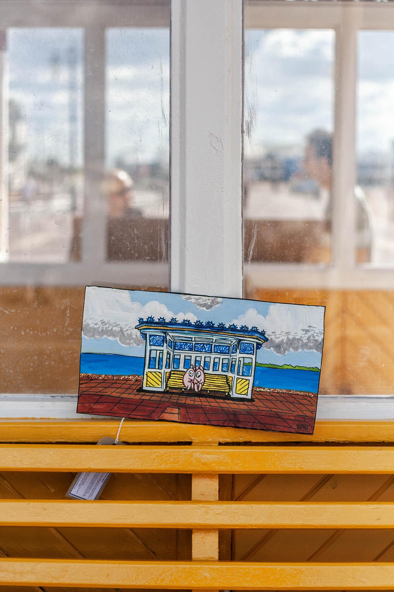 We're standing on a promenade in a seaside city, the pebbled beach somewhere to our right, a row of tall, Victorian or Edwardian seafront hotels and apartments somewhere to the left. We're close to a 19th century shelter set up on the promenade with iron benches painted yellow and arranged in an H-shape with the long bar running parallel to the sea. At the backs of each bench there are glass panes in painted white metal frames, all topped off with a metal canopy to protect people from the wind, rain, or sun, depending on the conditions. It's sunny today, with some bubbling cloud in the bright sky, and we can see that through the back of the shelter. We're close to one end with some bars of the yellow, metal bench seating taking up the bottom third of view, then two panes of glass above. Through those we can see to the opposite side of the H-shape where two people are turned towards one another talking, and beyond them, blurred, the promenade, the sky, and a hint of a pier stretching ou