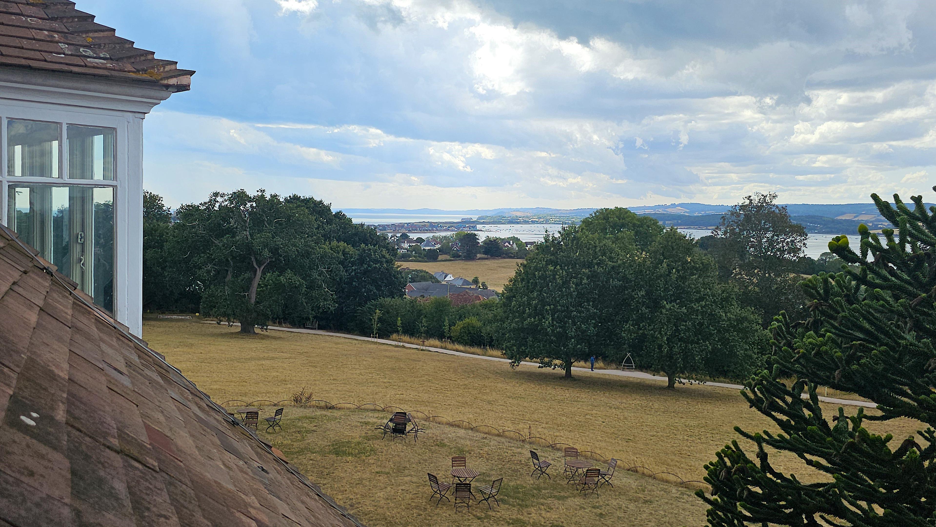 View from a first floor window over the Exe estuary. There is a red roof and a window to the left of the frame, with tables and chairs on a scorched lawn outside, and trees in the middle distance.