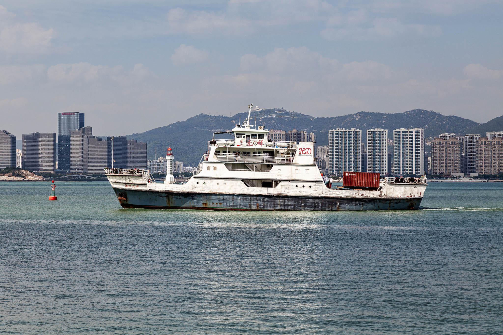 We're on one side of a wide stretch of calm water, looking across to a city skyline of skyscrapers on the distant shore backed by a more distant range of hills. It's a bright day with a little cloud and haziness in the air so the sky appears pale and the landscape over the water is similarly faint. Our focus is on a reasonably small car and passenger ferry passing to the left in the middle distance. The vessel has white upper decks over a hull that might once have been a deep blue but is now closer to a slate colour with lots of signs of neglect in the form of scratched paintwork and rust stains. The main decks are bunched in the centre of the ferry and there are four of them, tapering slightly towards a small bridge on the very top, and each with some open deck spots for passengers to enjoy the view. We can see a handful of people in gaps on the decks but more are gathered towards the aft behind a gap where the red container on a truck stands out for its colour.