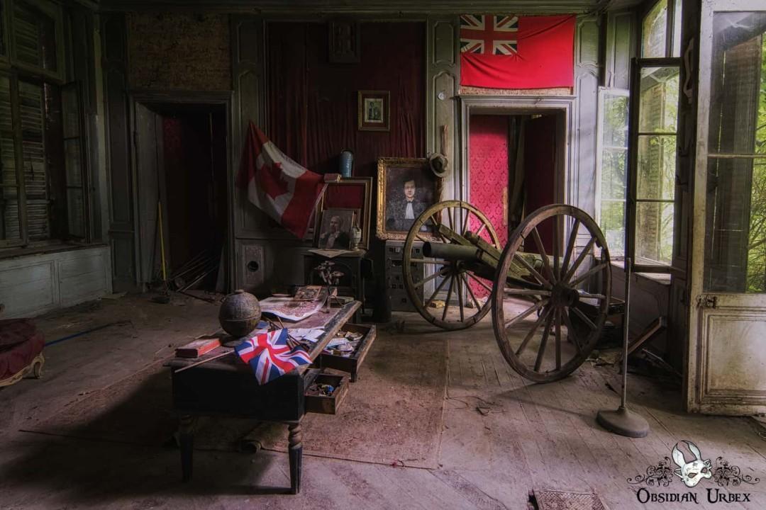 A derelict room in an abandoned chateau displays a cannon, Canadian flag, and a Union Jack and other interesting looking items