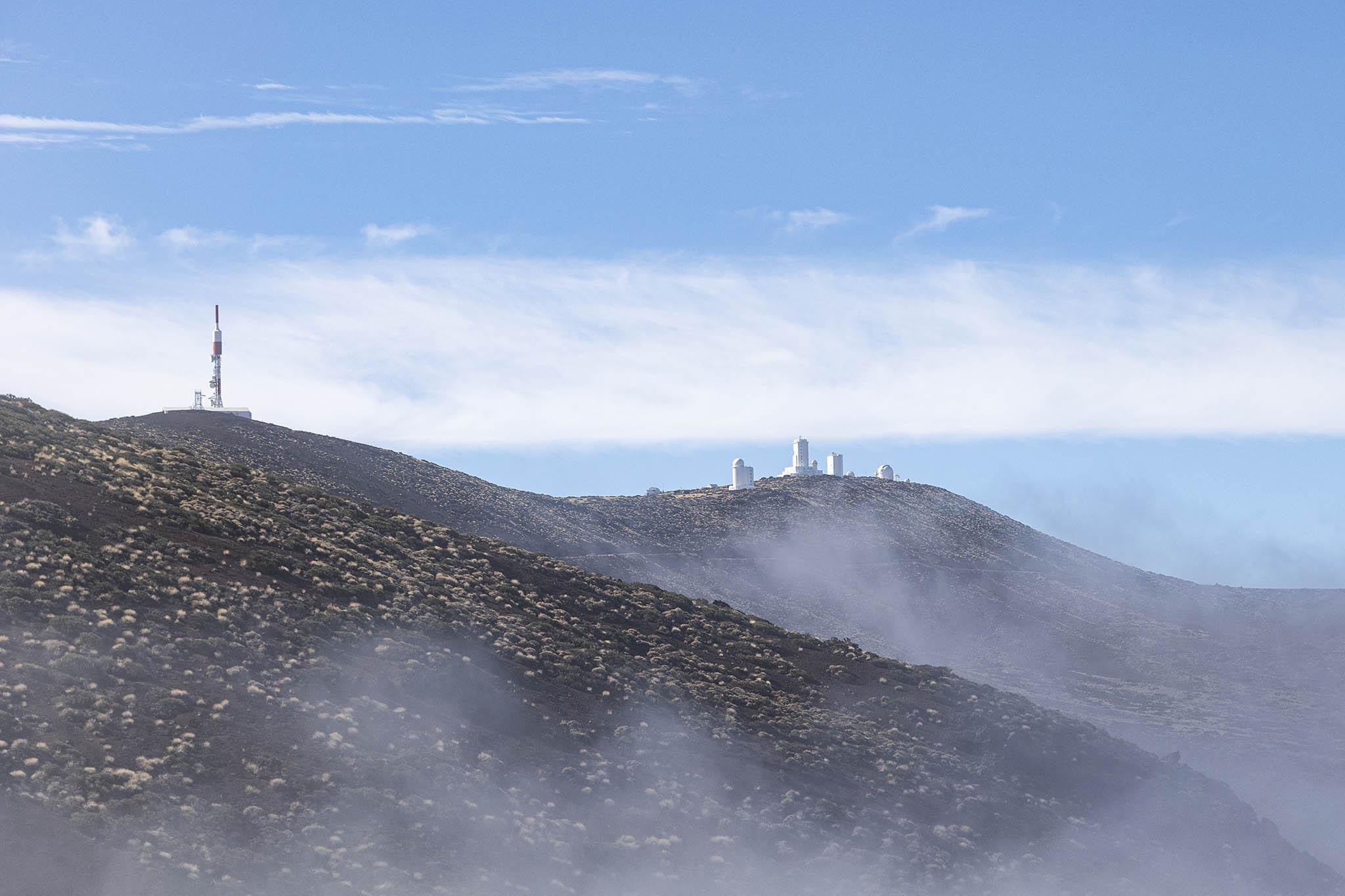 We're at high altitude on a volcanic, rocky surface with stony, hilly slopes overlapping ahead of us. Cloud low enough to touch the mountainous height we're at is just dissipating, leaving a mostly blue sky overhead and ghostly sheets of mist clinging to some of the hard ground. On a hill ridge a few hundred metres ahead we can see some white buildings. Some are rectangular but there are domes on a few which shows this to be a cluster of observatories. On the highest part of the curved ridge to the left there's a tall radio antenna. There's a coolness in the air from the height and the vanishing cloud cover.
