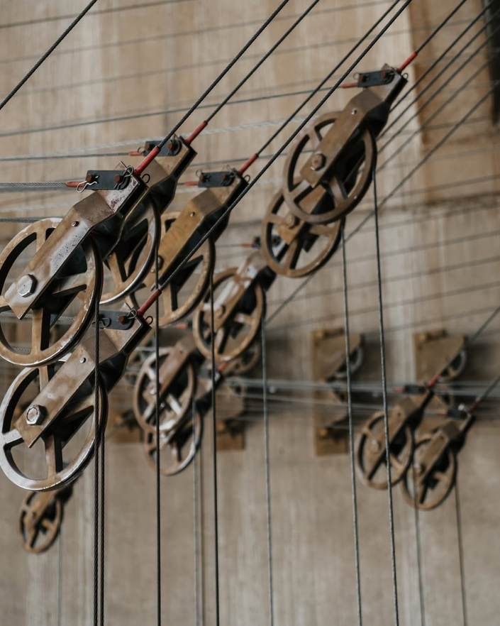 Color image of exposed gear and cable control mechanism for moving the acoustic ceiling panels in the circa 1973 E.J. Thomas Performing Arts Hall, The University of Akron, Akron, Summit County, Ohio, USA. Image shows multiple gears and pulleys attached to steel cables that form part of the mechanism for moving the acoustic ceiling panels inside the performance space of this 2,300 seat Brutalist-inspired performing arts hall, attached to and set against the backdrop of the concrete shell structure of the hall's soaring lobby area.