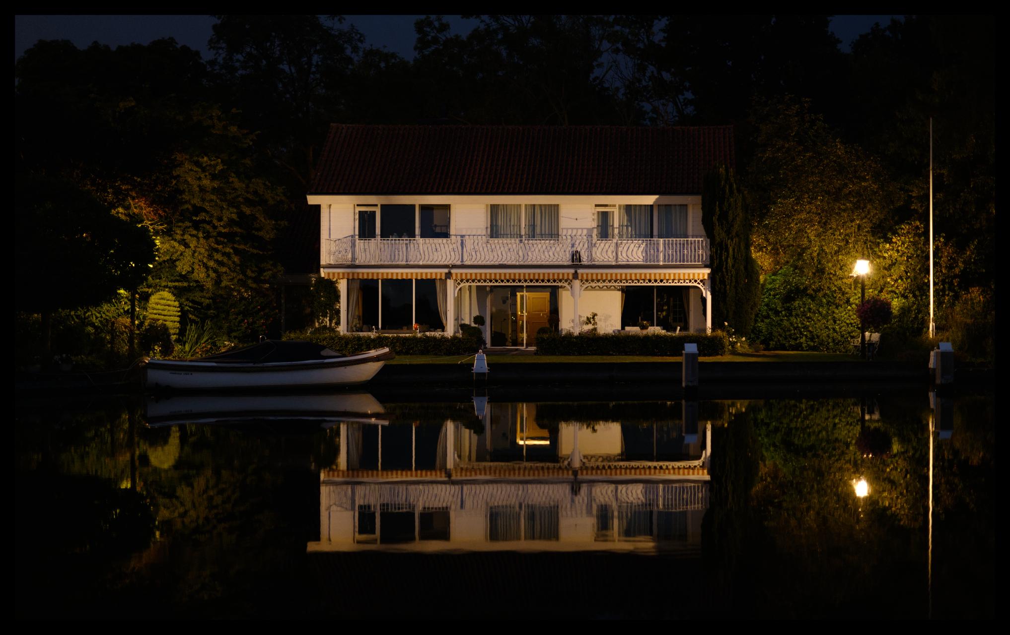 Night shot of a lit house on the waterfront. Mooring posts are visible and a boat is moored on the left. The whole scene is mirrored in the water. 

Another photograph from my ongoing 'Sleepless nights' series (#Nightlive).
 
PS: You've got to love image stabilisation in modern digital camera's: This was shot handheld using a full frame body with a 50mm lens at 1/4s. It being an almost windless night did help a bit......