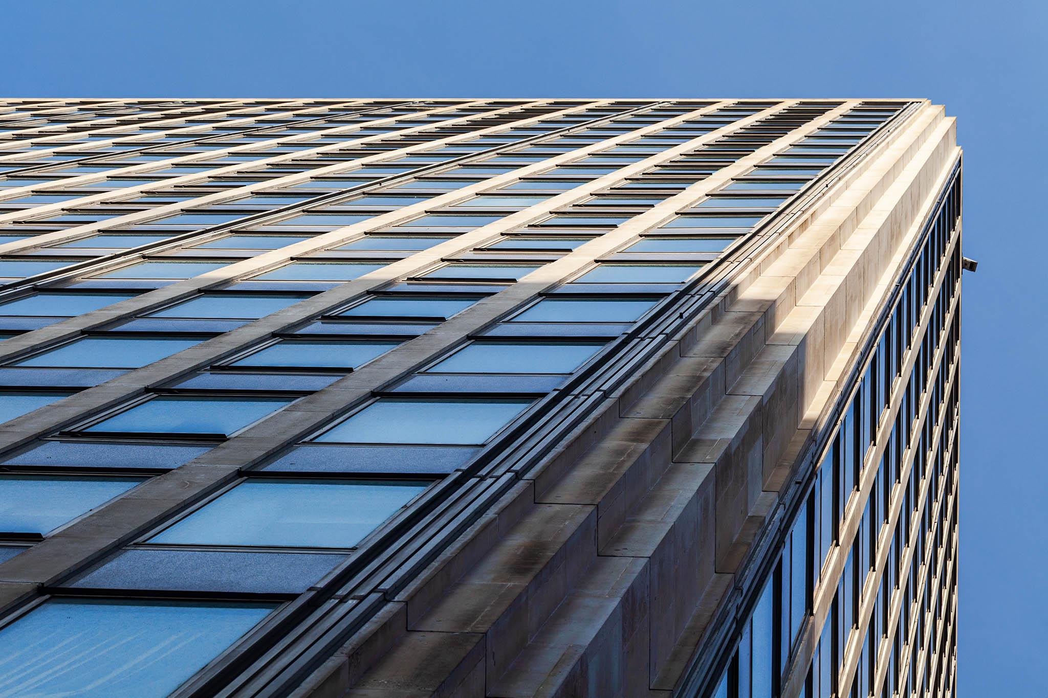 We're in a city street on a bright, cloudless day and we're looking up into the blue overhead. The sky forms a blue strip across the top and down the right side of view. The rest of the picture is taken up by a simple skyscraper block; it's not possible to tell if these are apartments or offices. The corner angles upwards from near the bottom left to the top right and is thick concrete. The two sides of the building we can see are repetitive columns of windows separated by thinner concrete strips. The lower half of the building - including where we're standing - is in shade while the sun catches the top half either side of the corner our eyes are drawn up.