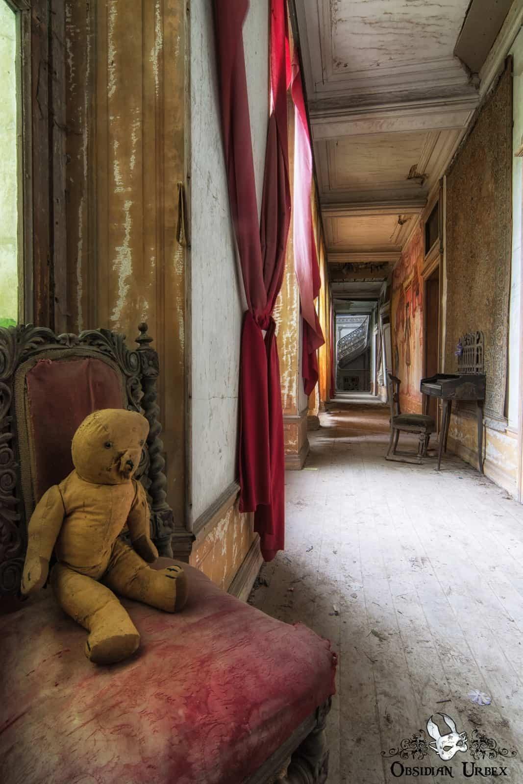 An abandoned chateau corridor features a faded yellow teddy bear on a red upholstered chair. Red drapes line the decaying hallway.