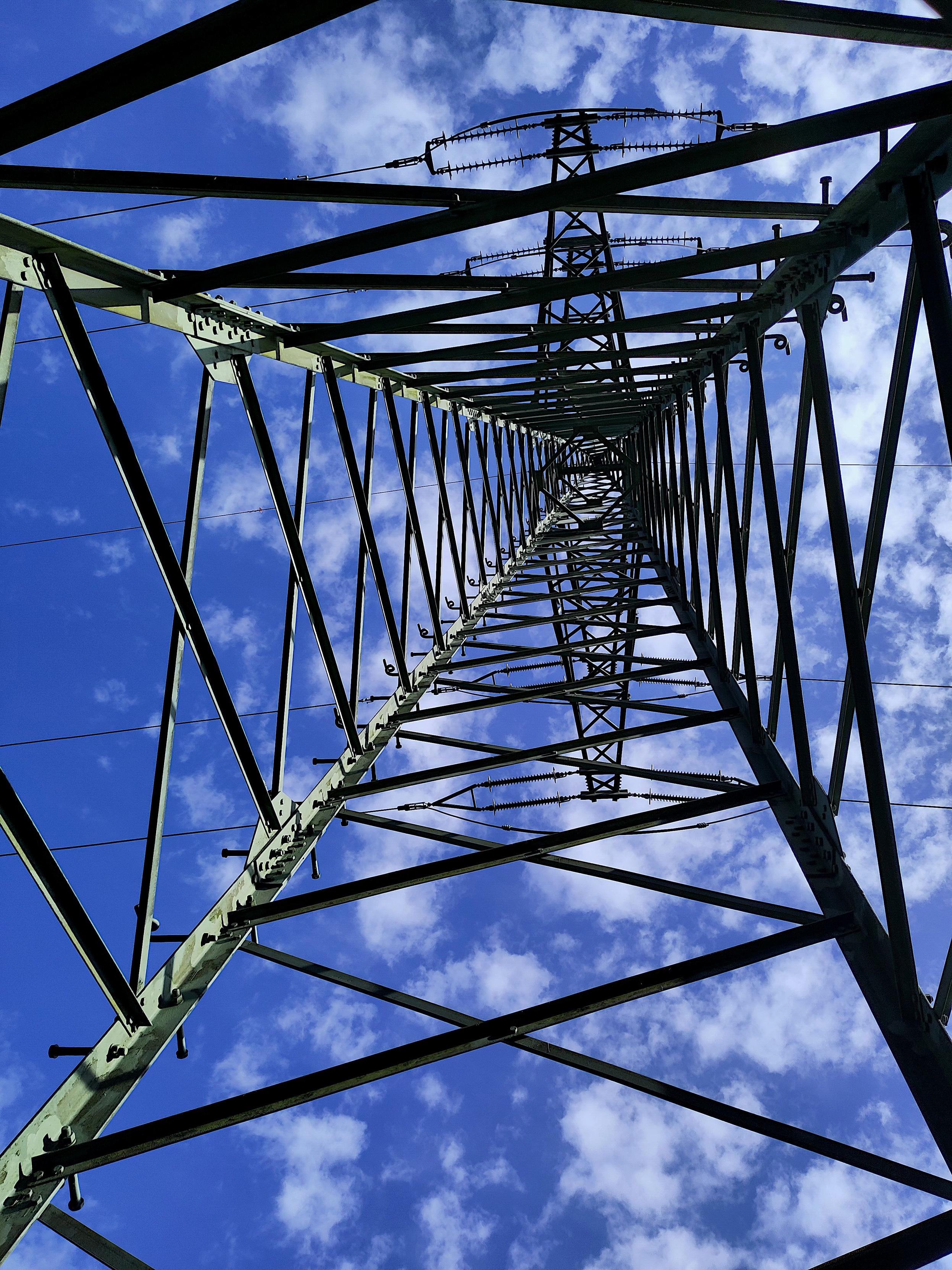 Blick senkrecht nach oben durch das Innere eines stählernen Gittermast-Strommasten, sodass die sich kreuzenden Streben und Querverbindungen symmetrisch zu einem kleinen Punkt hoch oben zusammenlaufen. Die Metallkonstruktion ist dunkel und als Silhouette zu sehen, mit Querträgern, Isolatoren und Stromleitungen im oberen Bereich. Der Hintergrund ist ein heller blauer Himmel mit kleinen weißen Wolken. Die Komposition ist symmetrisch und stark geometrisch.