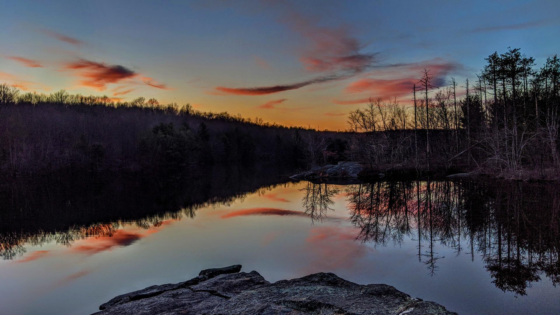 Standing on a dark gray rock outcrop a few feet above the shore of a lake in the deepening blue gray twilight of post sunset. The sky is filled with wispy swirls of cloud, tinted bright pink with dark purple shadows by the recently set sun. This is mirrored almost perfectly on the very slightly rippled lake surface. A forest of mostly leafless hardwoods with a few evergreens encircles the lake, mostly silhouetted by the dim evening light.