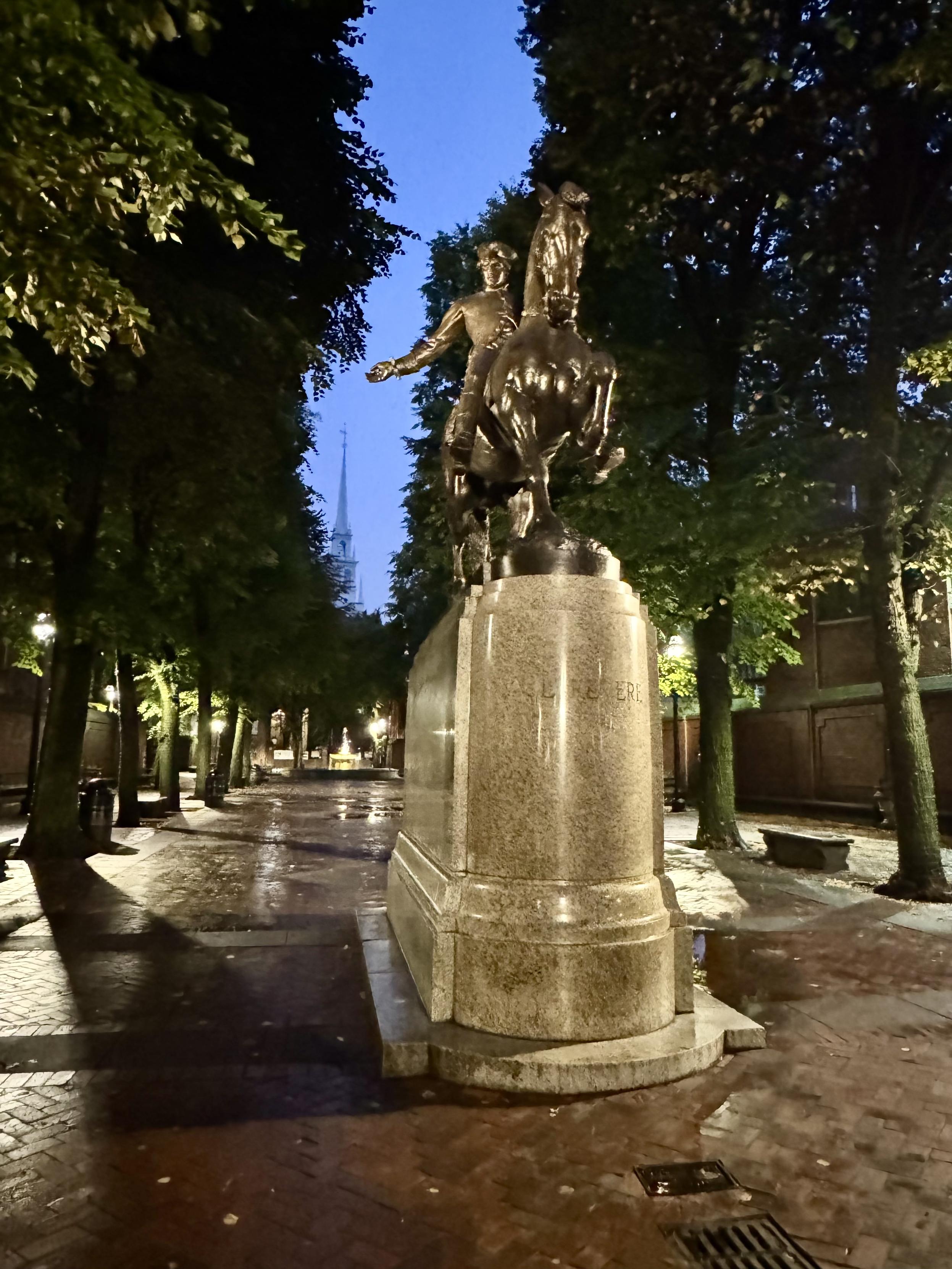 Photo taken at night of equestrian statue of Paul Revere, located in the Paul Revere Mall. The ground is covered with red bricks. The mall is lined with trees. The Old North Church is seen in the background.
