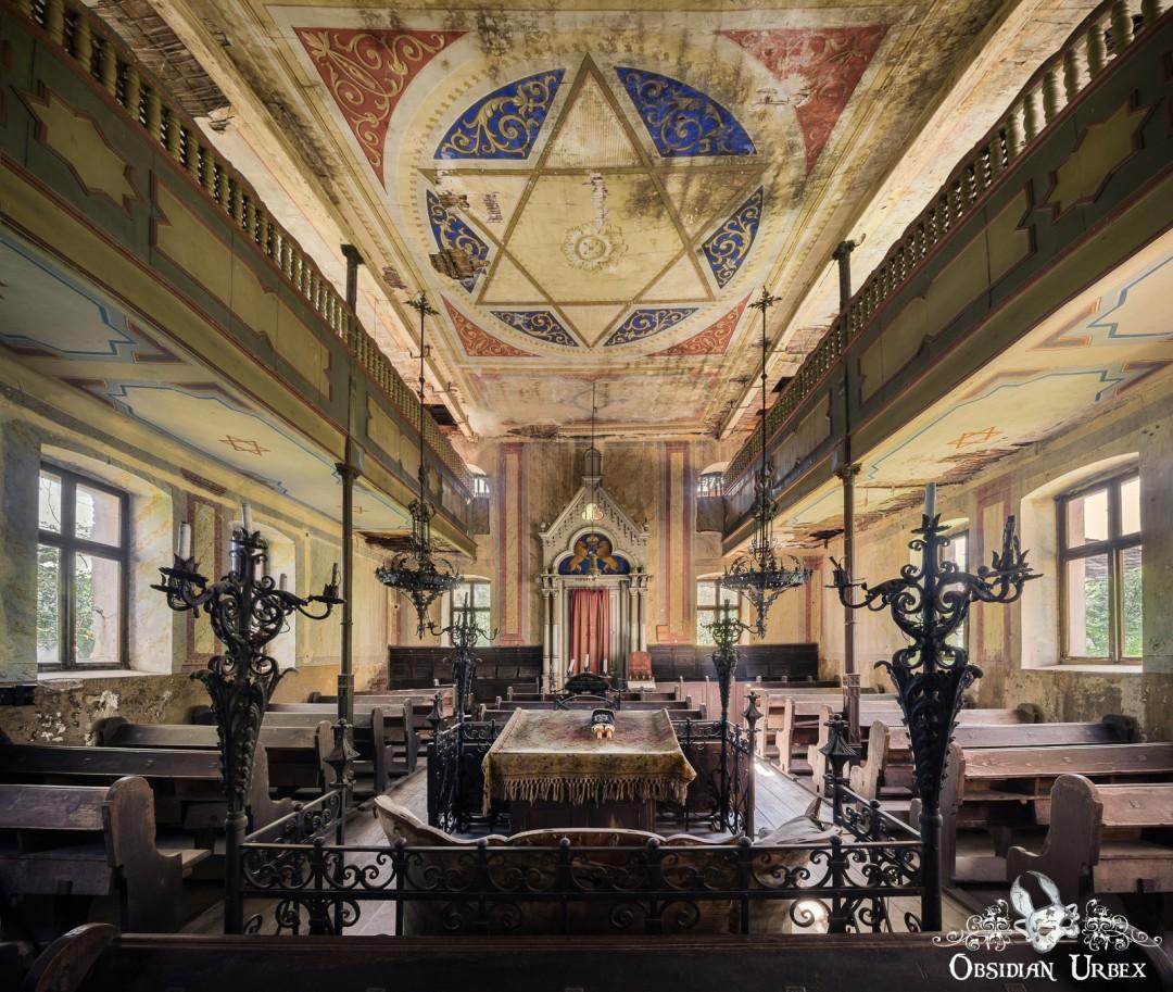 An ornate, decaying synagogue interior features a large Star of David on its painted ceiling. A bimah is central, surrounded by pews and balconies