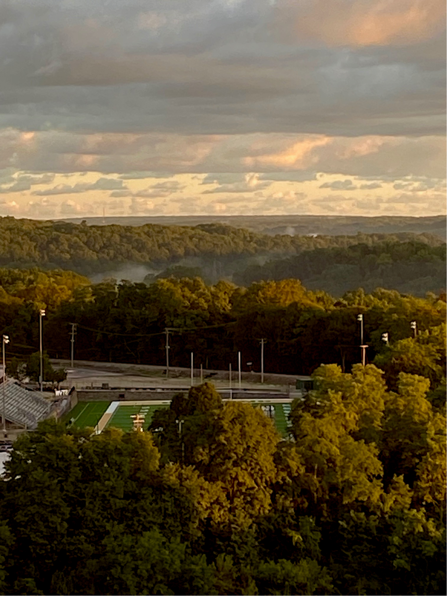 Color image of sunrise 14 September 2026 over Akron, Summit County, Ohio, USA. Image shows the sun lighting the Cuyahoga River Valley beyond the St. Vincent-St. Mary High School stadium on Green Street, with trees in the lower part of the photo, the stadium center with lights and stands on both sides, then more trees with mist and fog rising from the river below, more distant trees along the horizon, and morning cumulus clouds in white, orange, yellow, gray and blue, giving way to patches of blue sky.