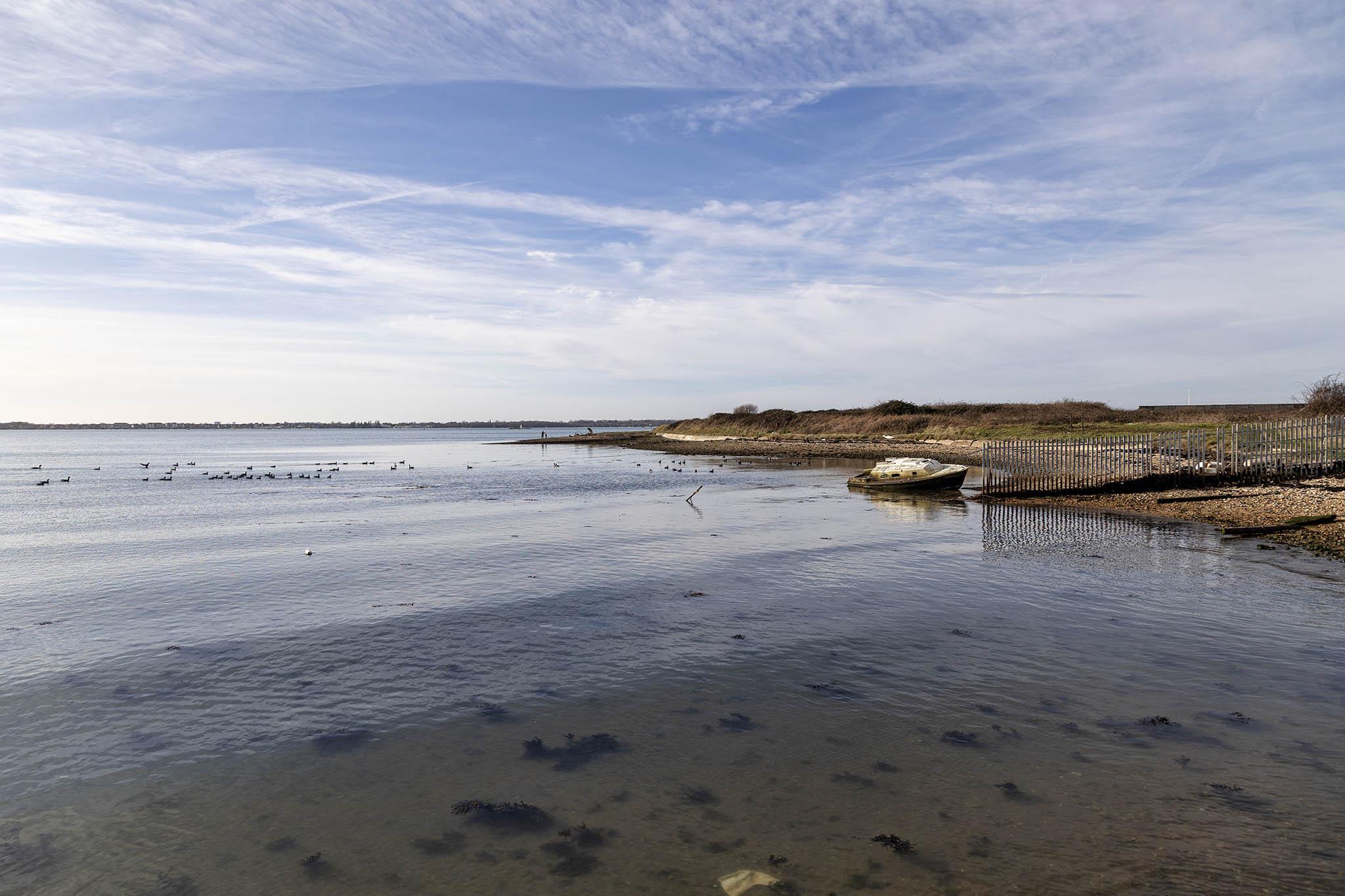 A tranquil scene set beside flat water and low land on a day with plenty of blue sky visible between the thin, almost transparent sheets and patches of white cloud overhead. We're next to a wide harbour but all the infrastructure and vessels associated with it are out of shot somewhere far to the left. We're looking out over the harbour water which has only the faintest of surface colouration differences to suggest there's any sort of tidal action here. The water is clear and very shallow near us and we can make out small clumps of black seaweed over a dark, sandy surface. A tiny strip of darkness on the horizon marks the far side of the harbour. On the right of us there's more land jutting outwards, reaching about halfway into the scene at its furthest point. The land is low with a shallow shingle shoreline that reaches back several metres until it hits grass that rises a little and is then lined with bushes that appear dry. There's a metal fence that separates off part of the land fr