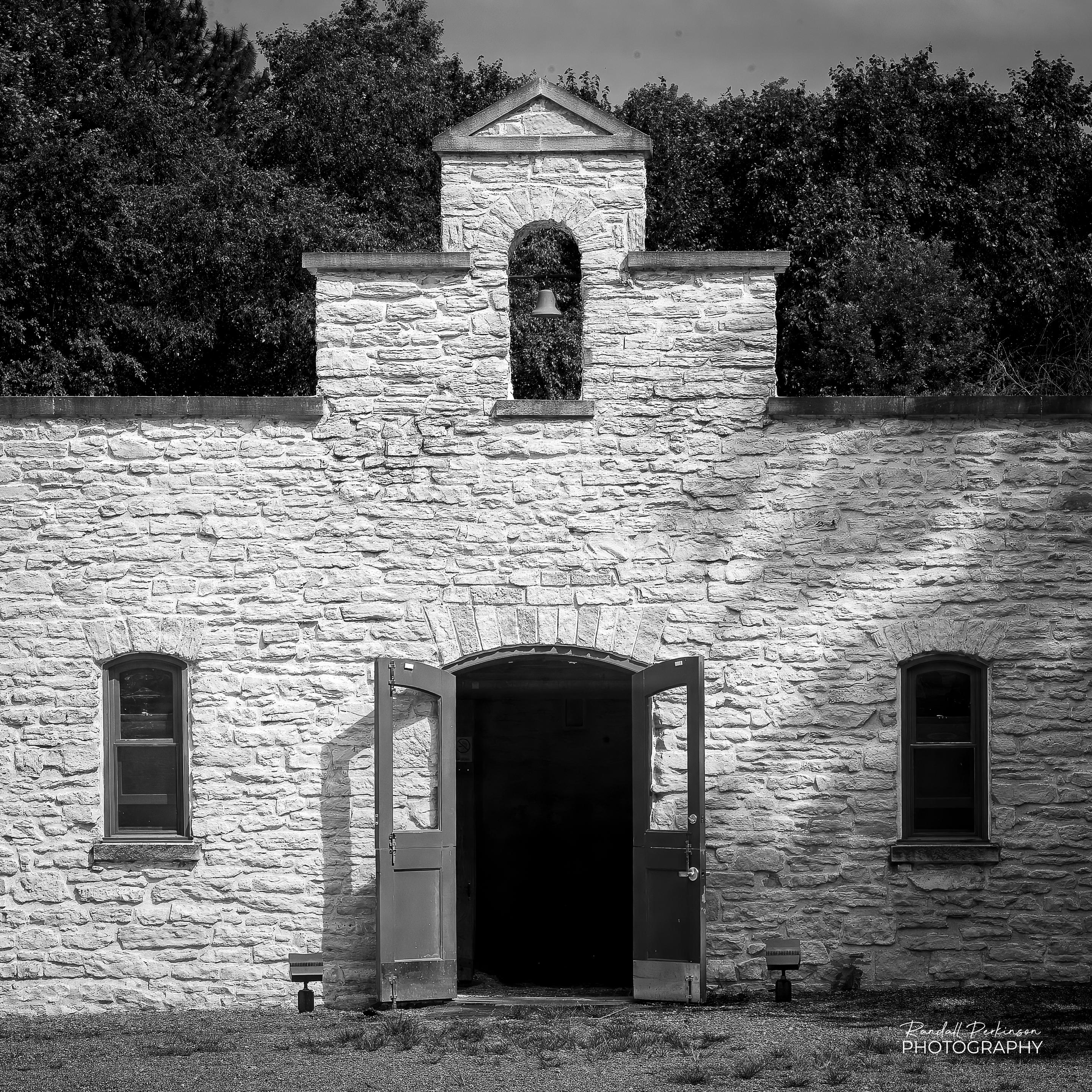 The front of a stone building with an open, double arched door at the center, a small arched window on either side of the door.  The front face of the building rises up at he center and provides an opening with a bell.