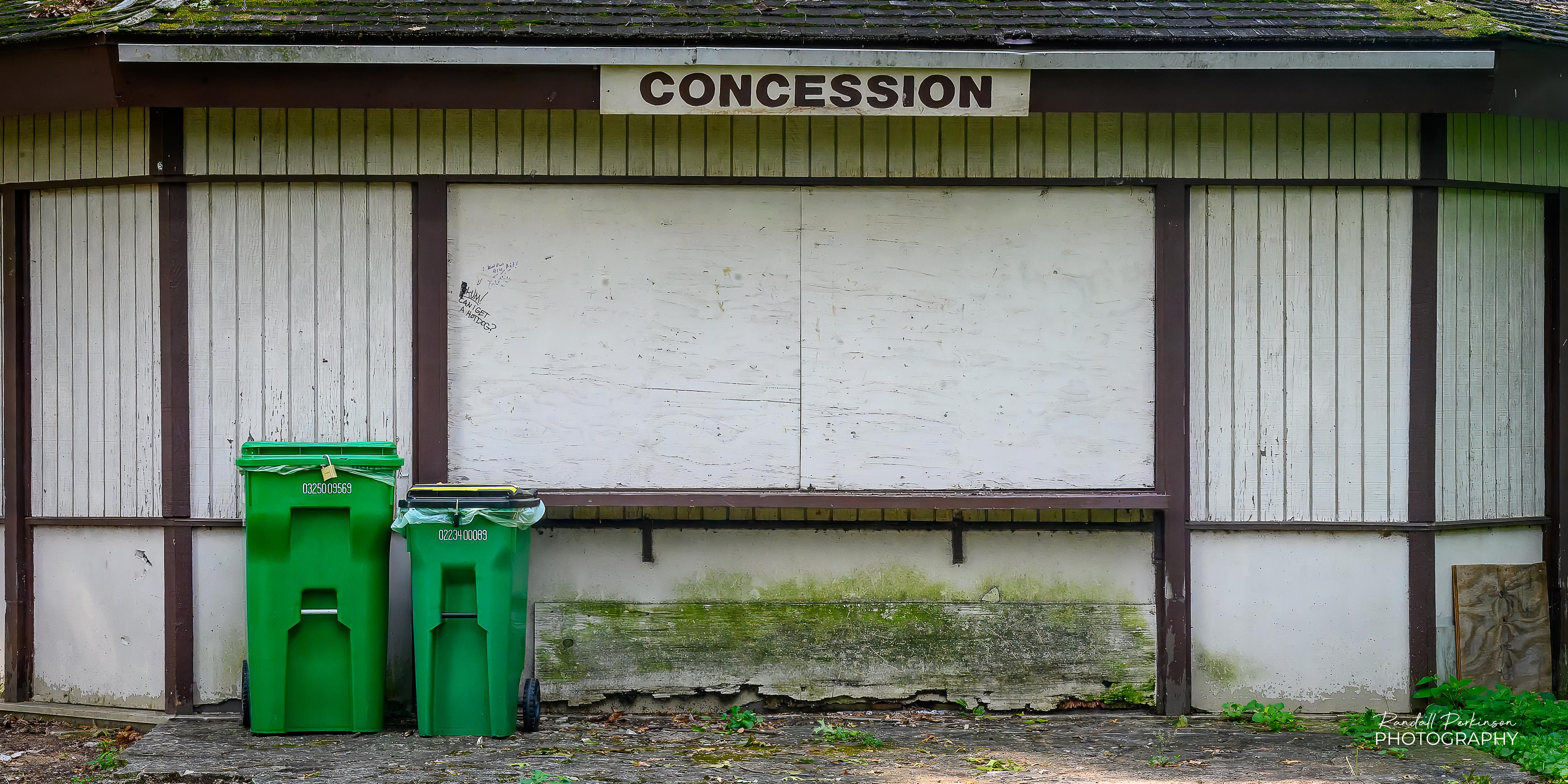 The front of a one-story wooden building with decaying fascia boards.  There is a sign reading "CONCESSION" over a wide opening that is boarded up.  There is a large and small green trash cart sitting in front of the building.