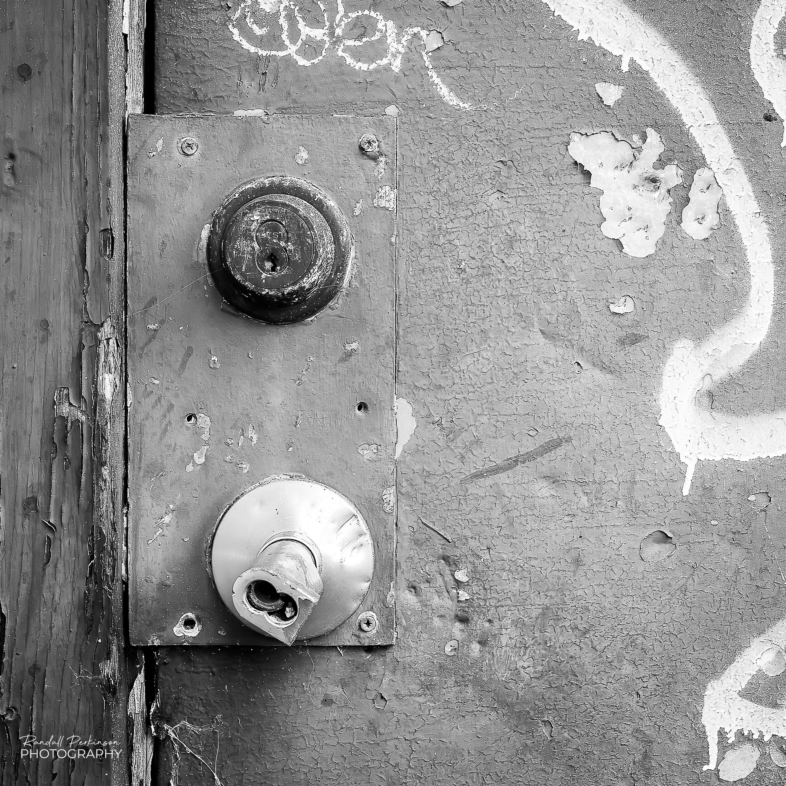 Close up image of a door with graffiti, a broken off door handle, and a rusty lock.