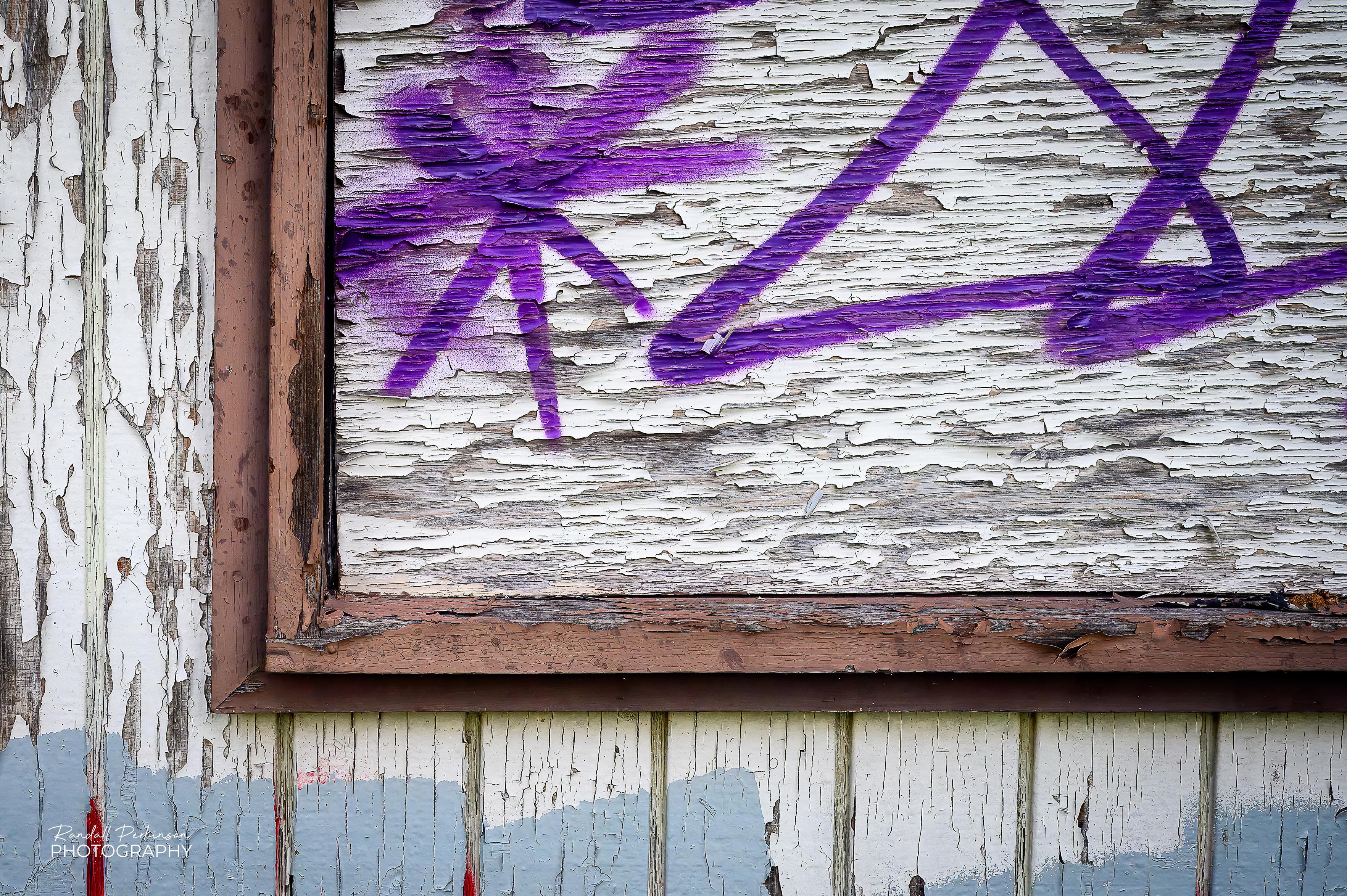 A closeup of one corner of a boarded-up window showing some purple spray-painted graffiti and the weathered, peeling paint.