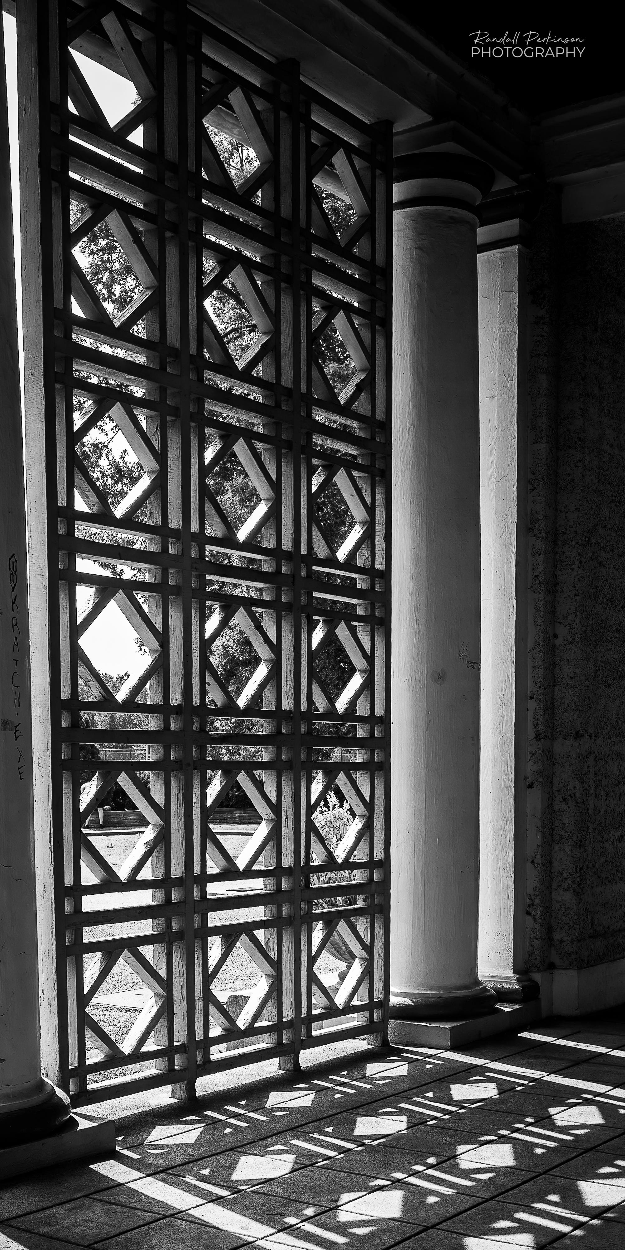 Light shines through a geometrically patterned trellis of a park building casting a detailed shadow on the floor inside the building.