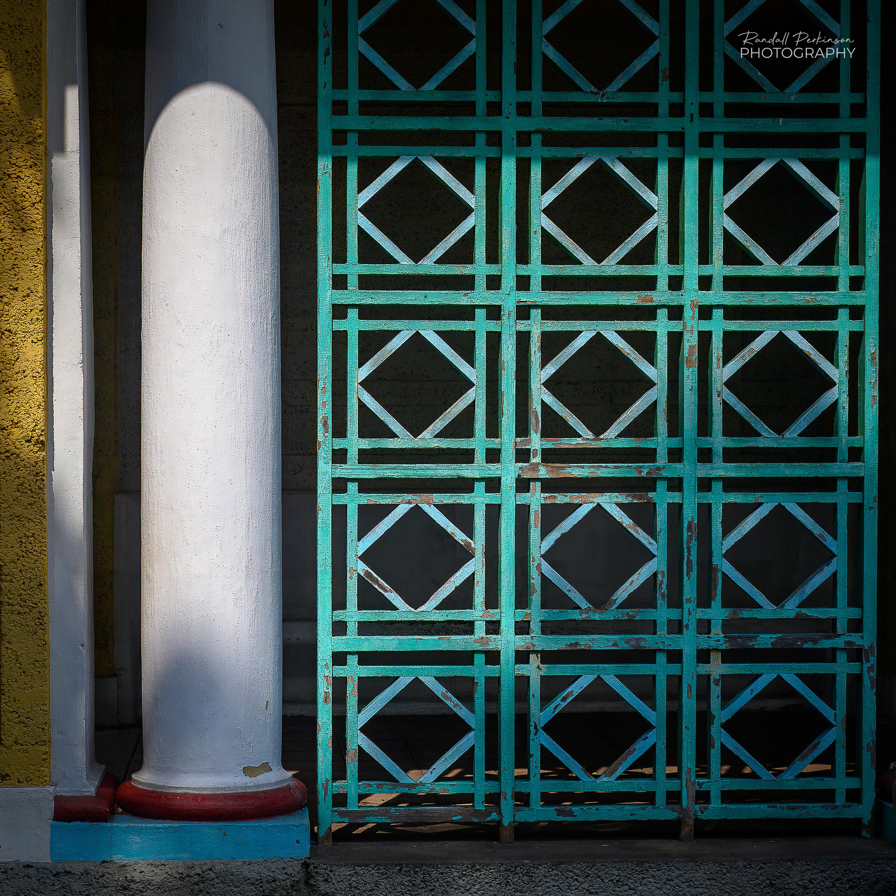 A green wooden trellis with multiple rows of vertical and horizontal boards forming squares filled with diamonds is next to a round column on the exterior of a park building.