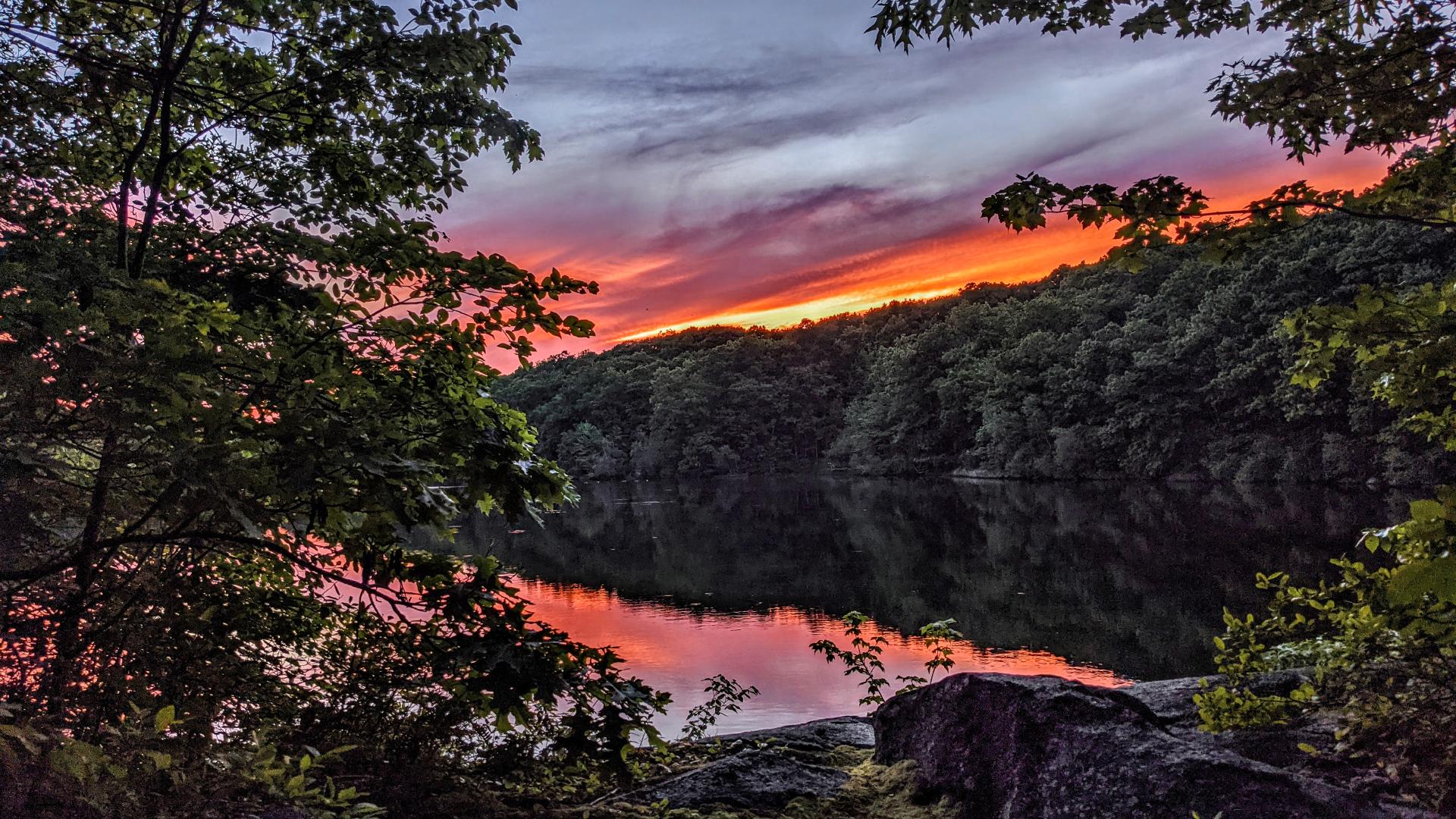 We are standing beside a pond at a small rocky, mossy break in the surrounding lush green forest. The evening sky is mostly cloudy, with a slim break just along the horizon filled with bright gold and orange sunlight. That break is allowing the recently set sun to tint the bottom of the clouds near the horizon dark pink, almost red. The rest of the clouds are gray with a slight violet tinge.