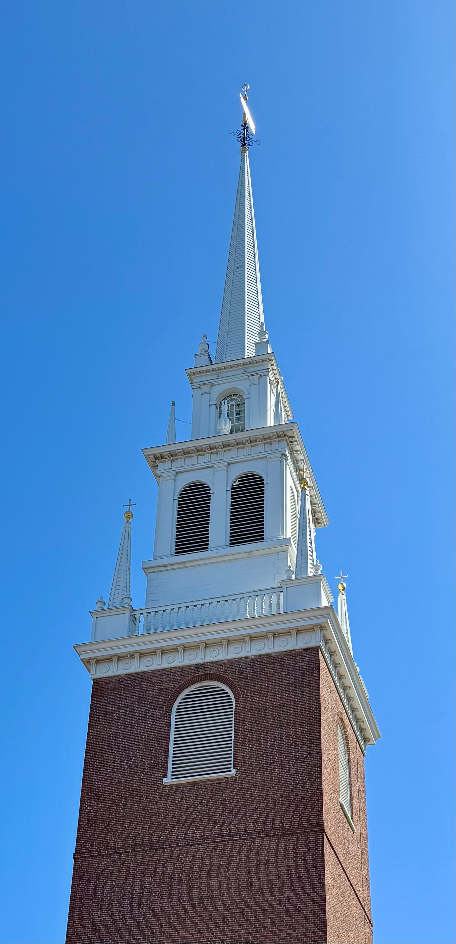 Photo of church steeple as viewed looking up from ground.  The base is red brick and the top is intricate white woodwork.  The top of the pointed steeple has a brass weathervane that is reflecting the bright sun.  Solid blue sky overhead.

This is the famous "Old North Church".  This is the church where lanterns were claimed to have been hung in the steeple (under the direction of Paul Revere) to signal the arrival of British troops in 1775--- "one if by land, two if by sea".  Here, "sea" was the nearby Charles River.  This image is the front of the church--- its backside can be seen in the background in the previous post in this thread.