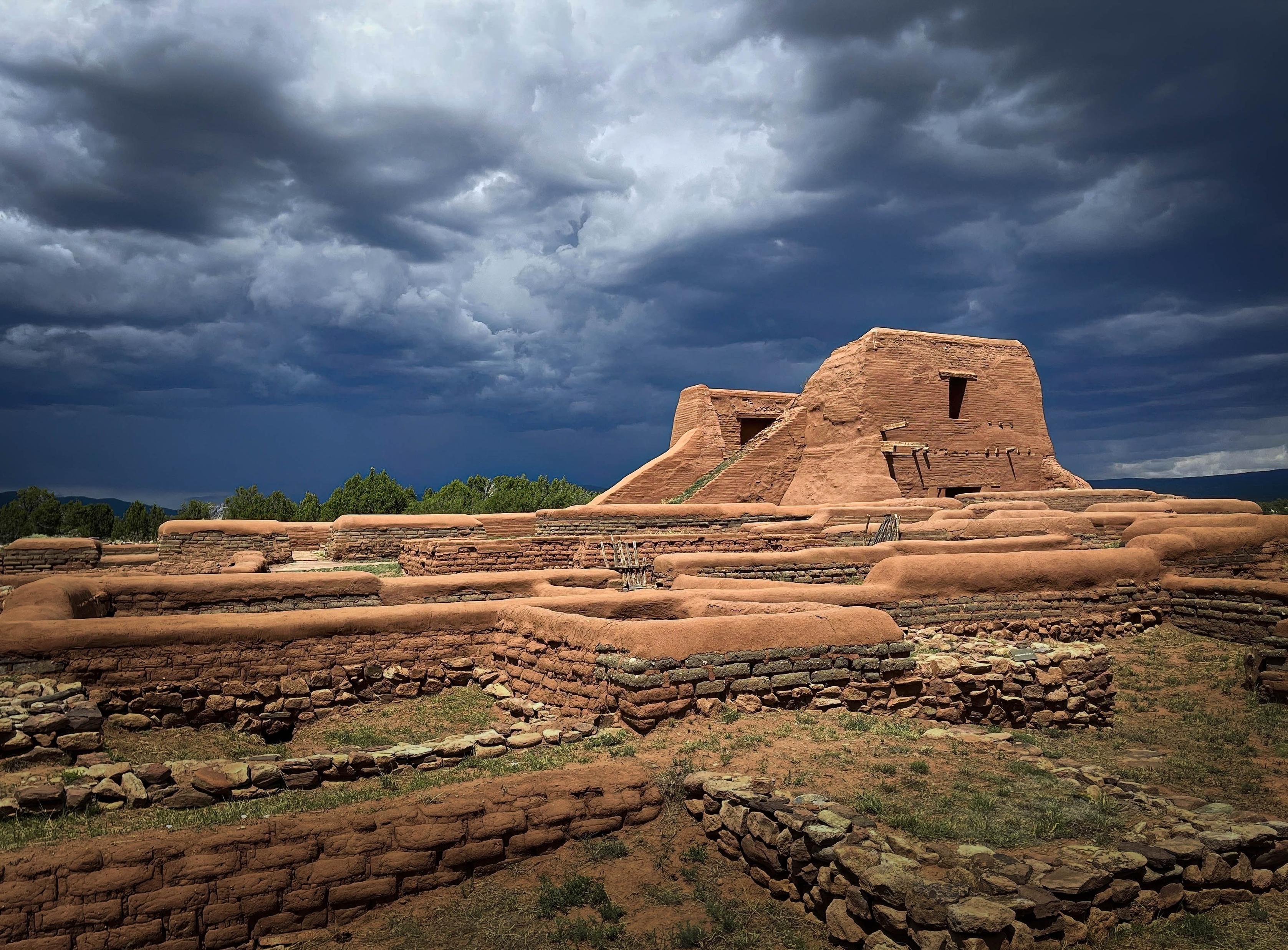 Adobe ruins at Pecos National Historical Park in New Mexico glow in warm sunlight beneath a dramatic sky filled with massive dark blue and gray thunderclouds. Low stone and adobe walls fill the foreground, with the larger adobe mission ruins rising against the storm in the distance.