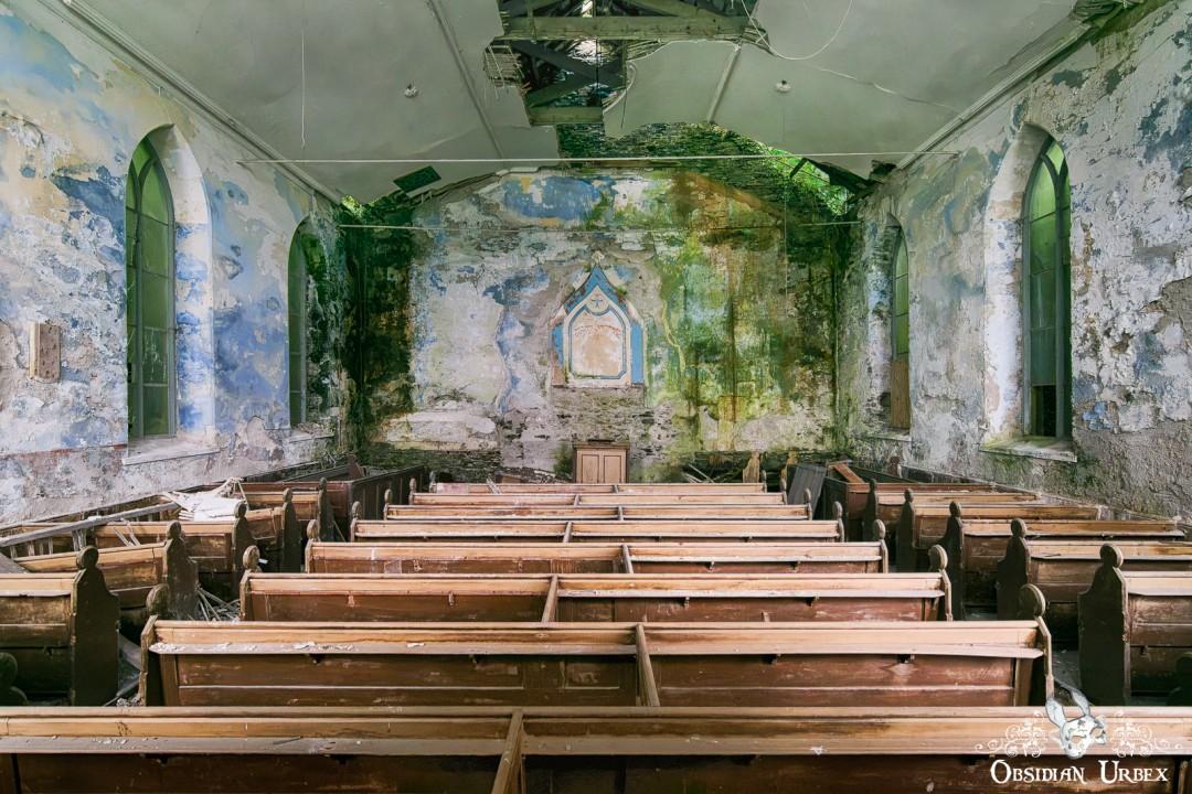 An abandoned church interior with rows of wooden pews leading to a moss-covered altar. Peeling paint and natural decay are visible throughout the space.