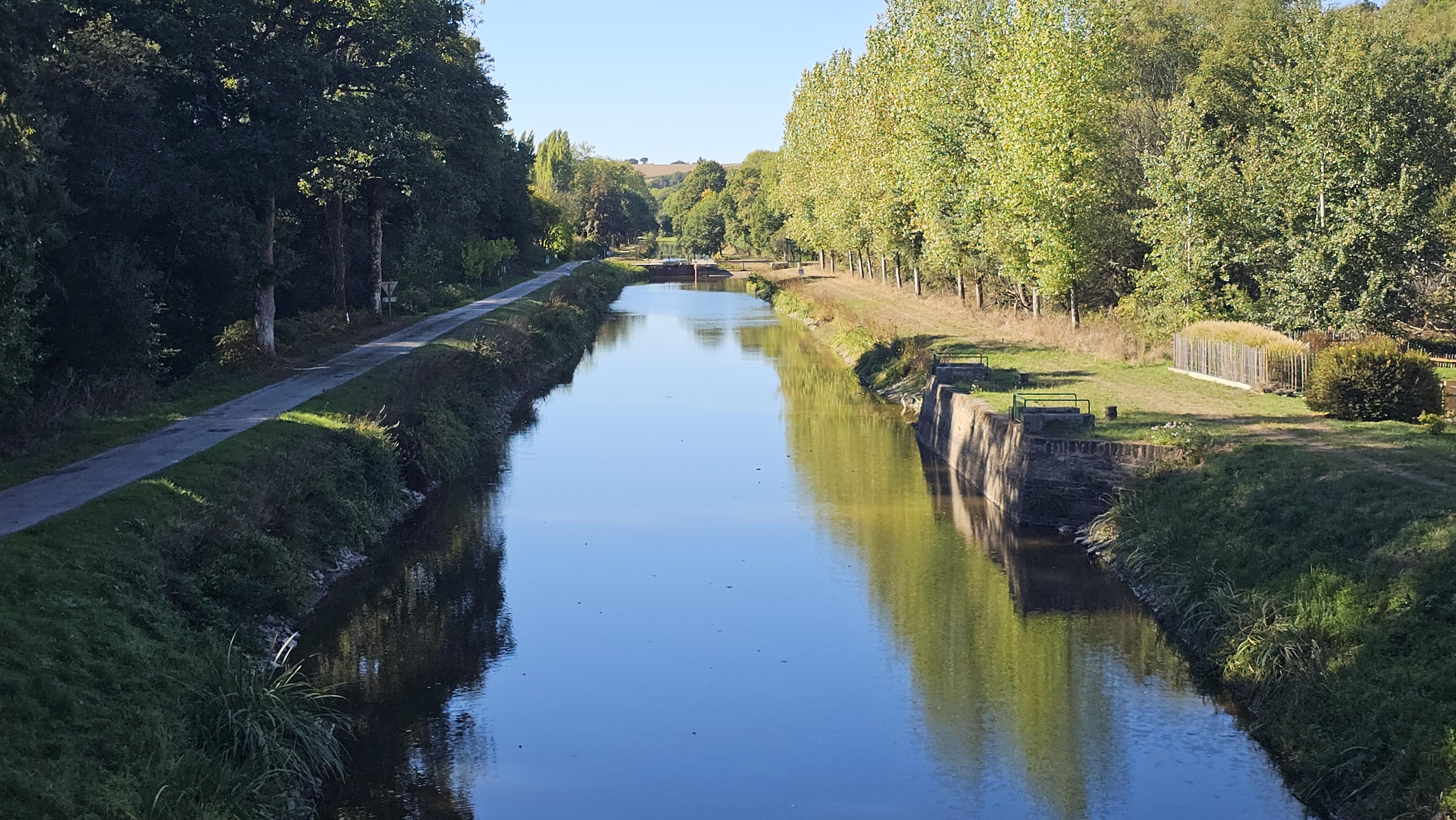 View along a canal, in sunshine, with trees either side and a towpath to the left.